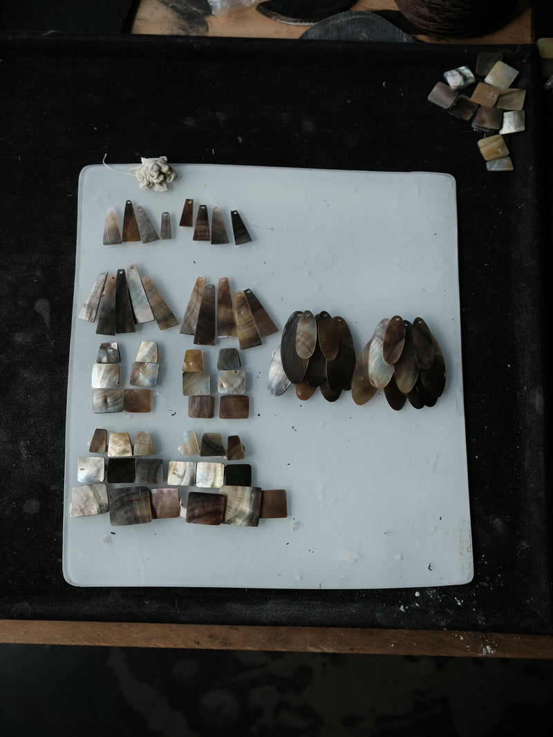 A collection of handmade jewelry components, including various cone-shaped and rectangular pieces made from natural mother-of-pearl and brown stone, neatly arranged on a white cutting board.