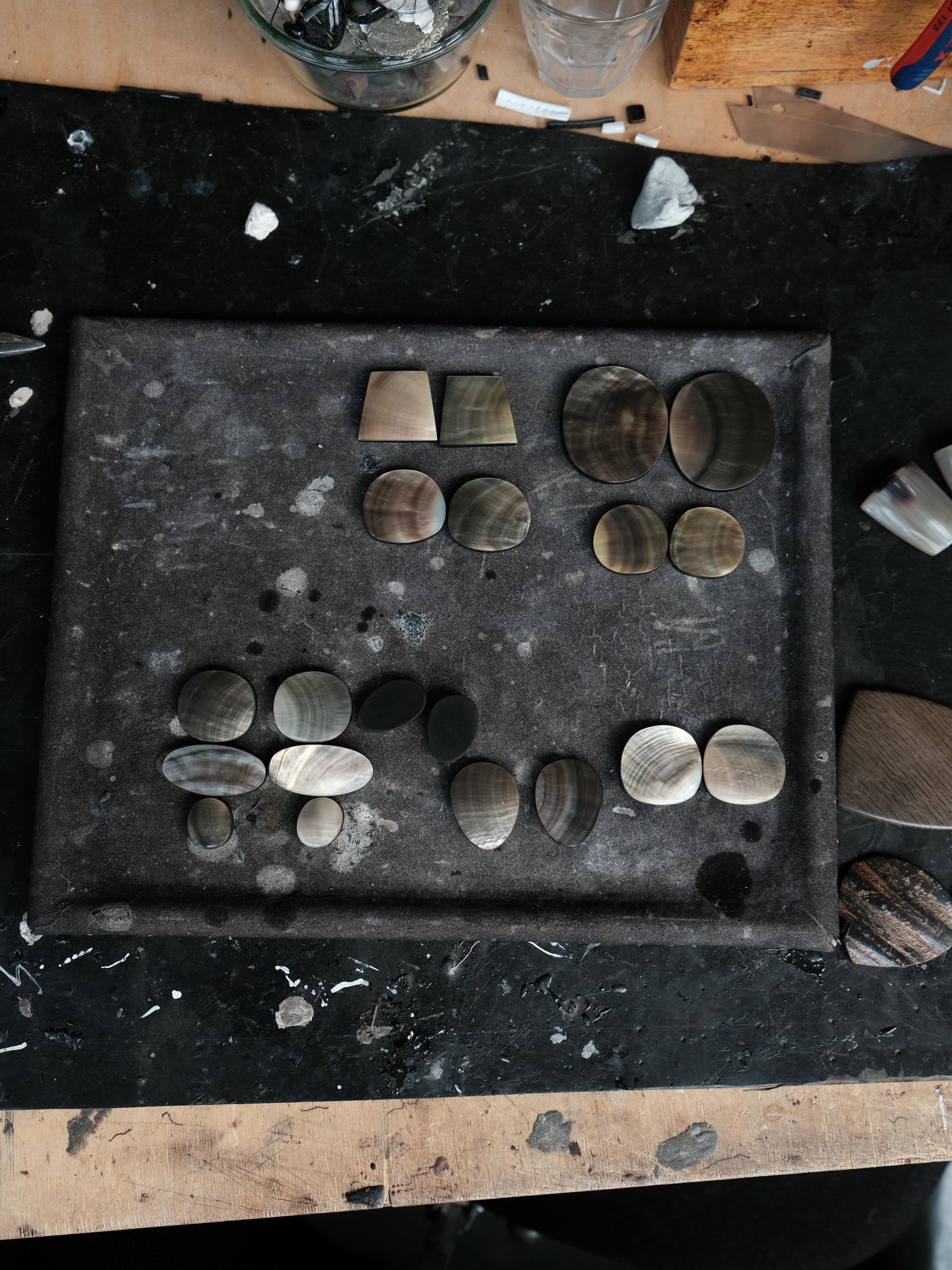 A dark metal tray holding multiple polished stone or shell-like pieces in varied oval, round, and geometric shapes, photographed from above on a worn black workbench with scattered debris and tools around the edges.