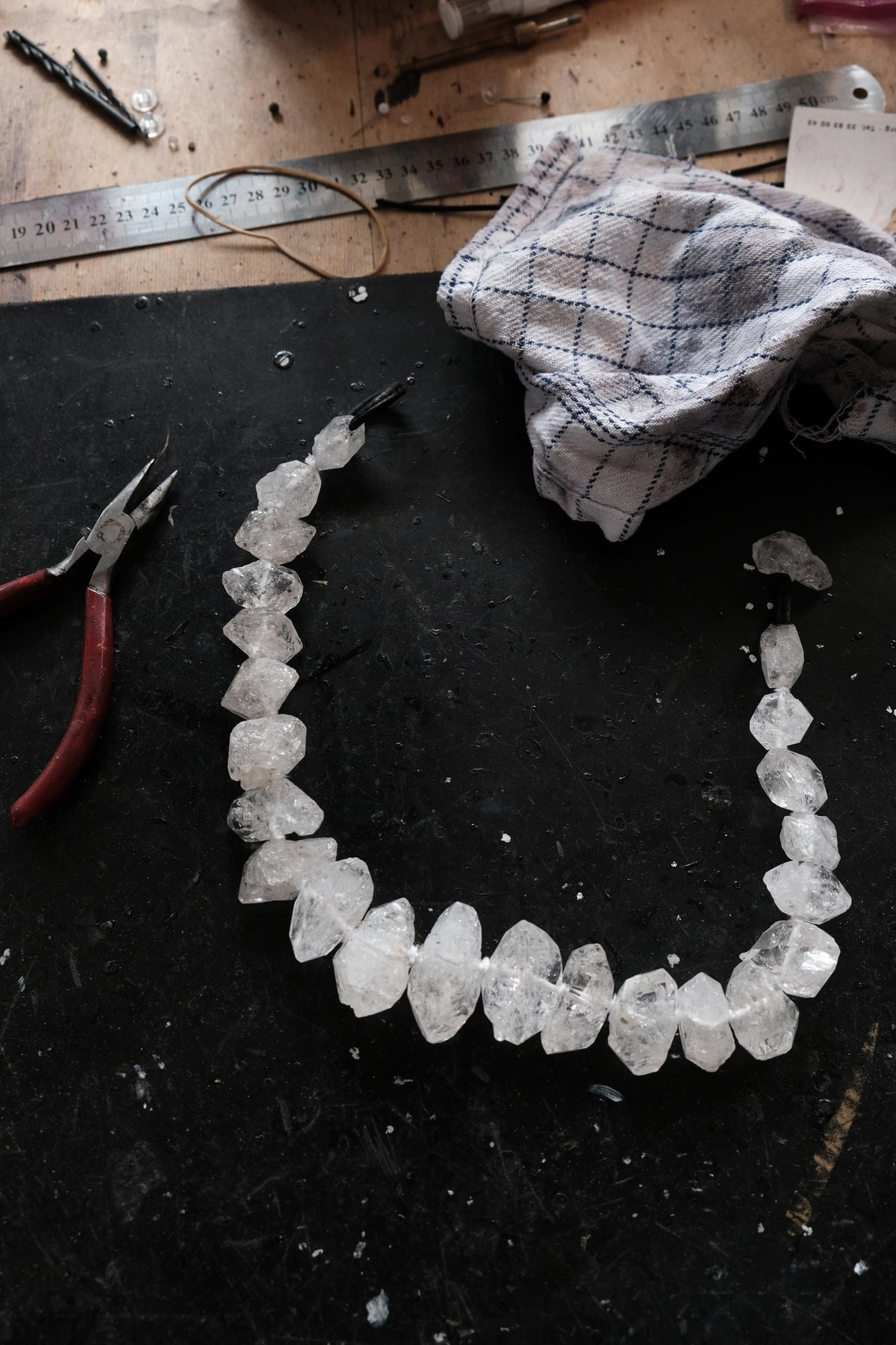 A raw crystal bead necklace made of irregular clear quartz stones, laid out on a dark workbench with pliers, a metal ruler, and a cloth nearby.