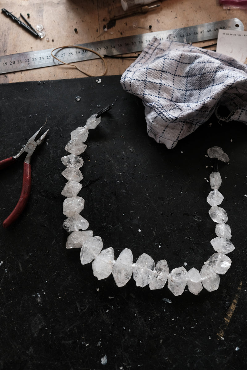 A raw crystal bead necklace made of irregular clear quartz stones, laid out on a dark workbench with pliers, a metal ruler, and a cloth nearby.