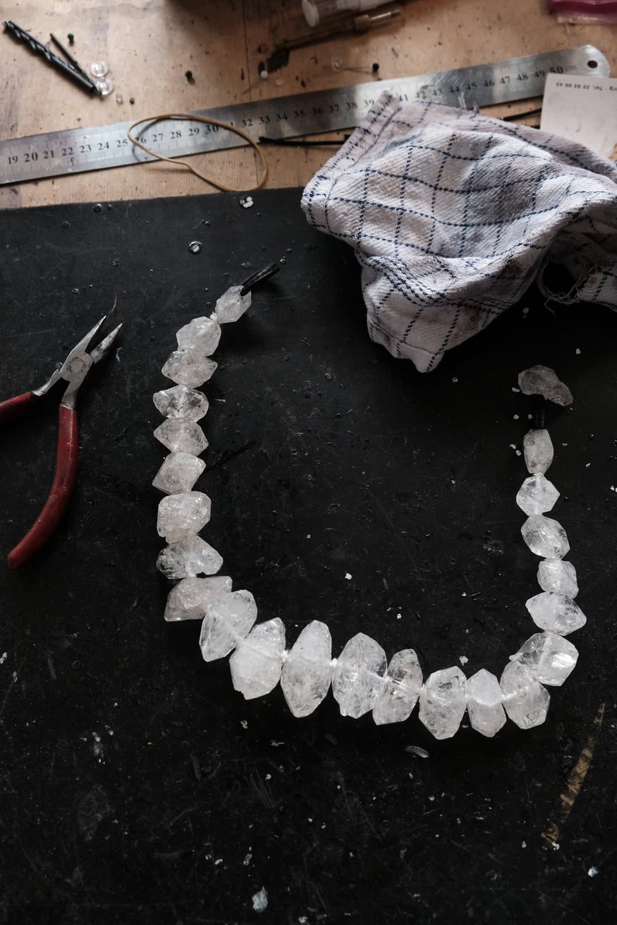 A raw crystal bead necklace made of irregular clear quartz stones, laid out on a dark workbench with pliers, a metal ruler, and a cloth nearby.