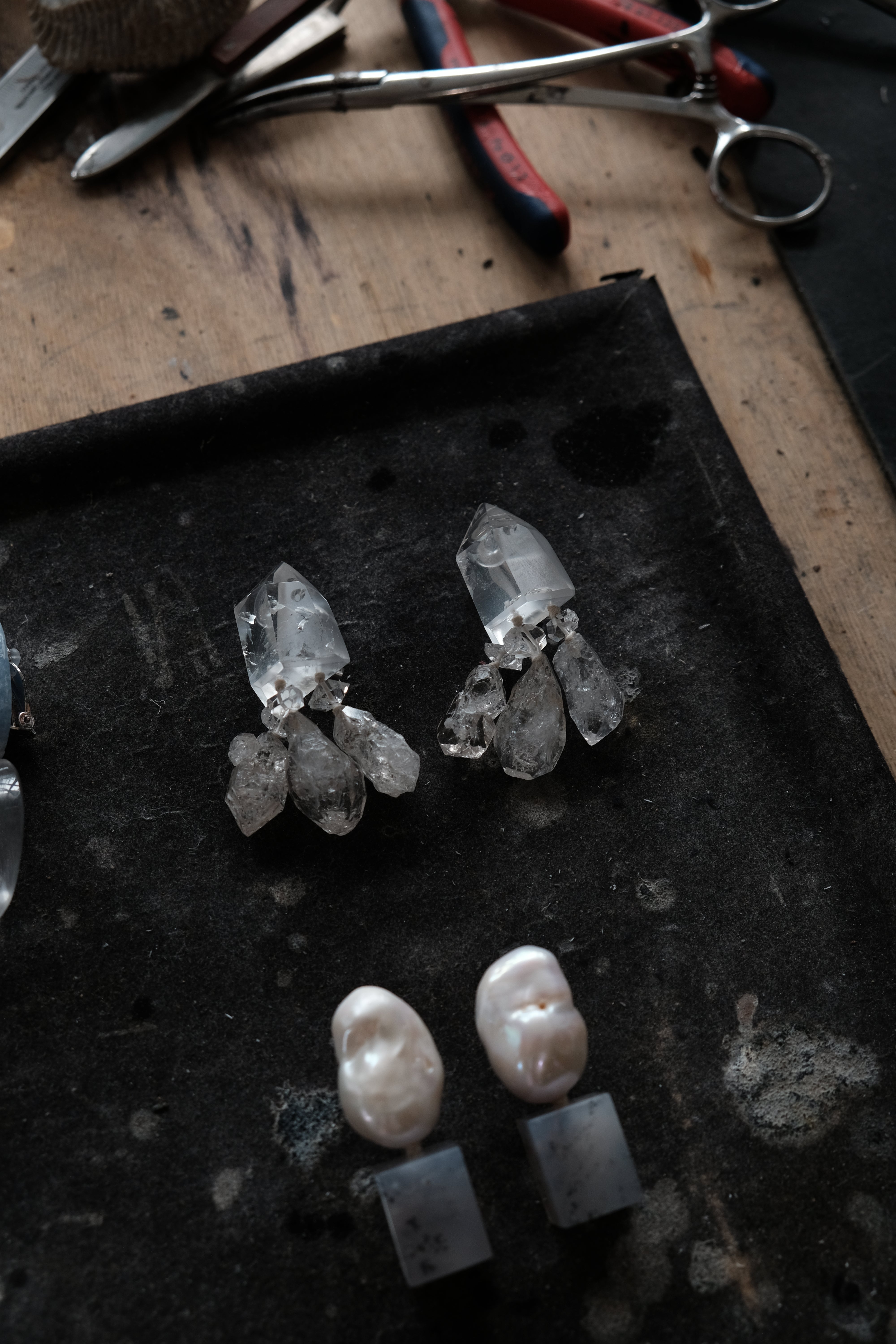 A pair of handmade crystal and pearl stud earrings displayed on a dark tray, with metal pliers and tools visible in the background on a wooden workbench.