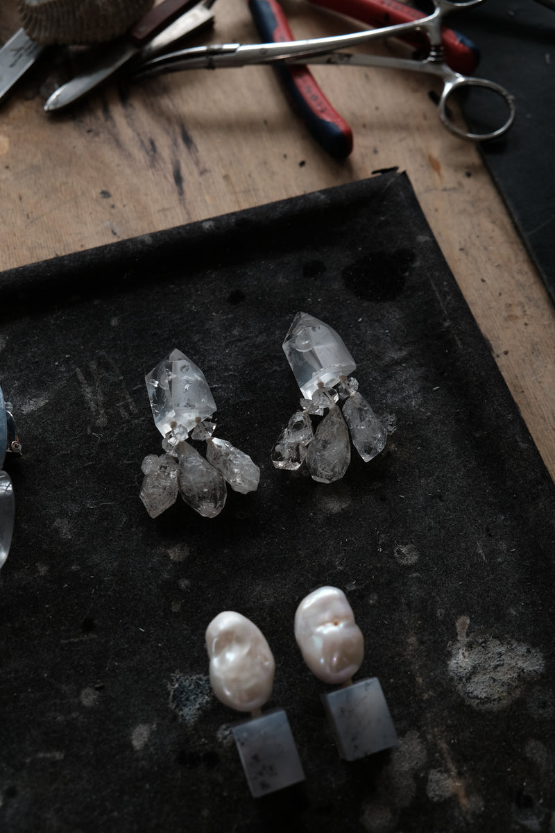 A pair of handmade crystal and pearl stud earrings displayed on a dark tray, with metal pliers and tools visible in the background on a wooden workbench.
