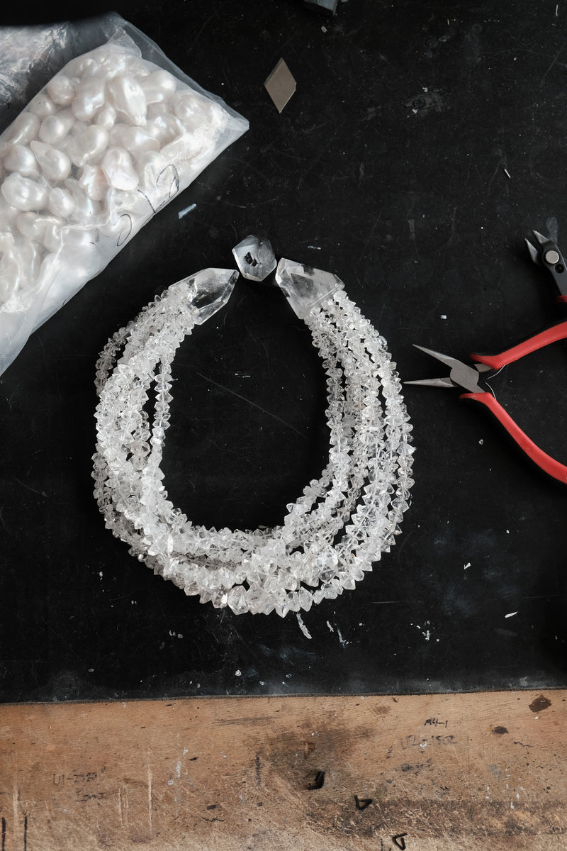 A handmade multi-strand necklace made of clear faceted crystals, displayed on a black workbench alongside a bag of white crystals and red-handled pliers.