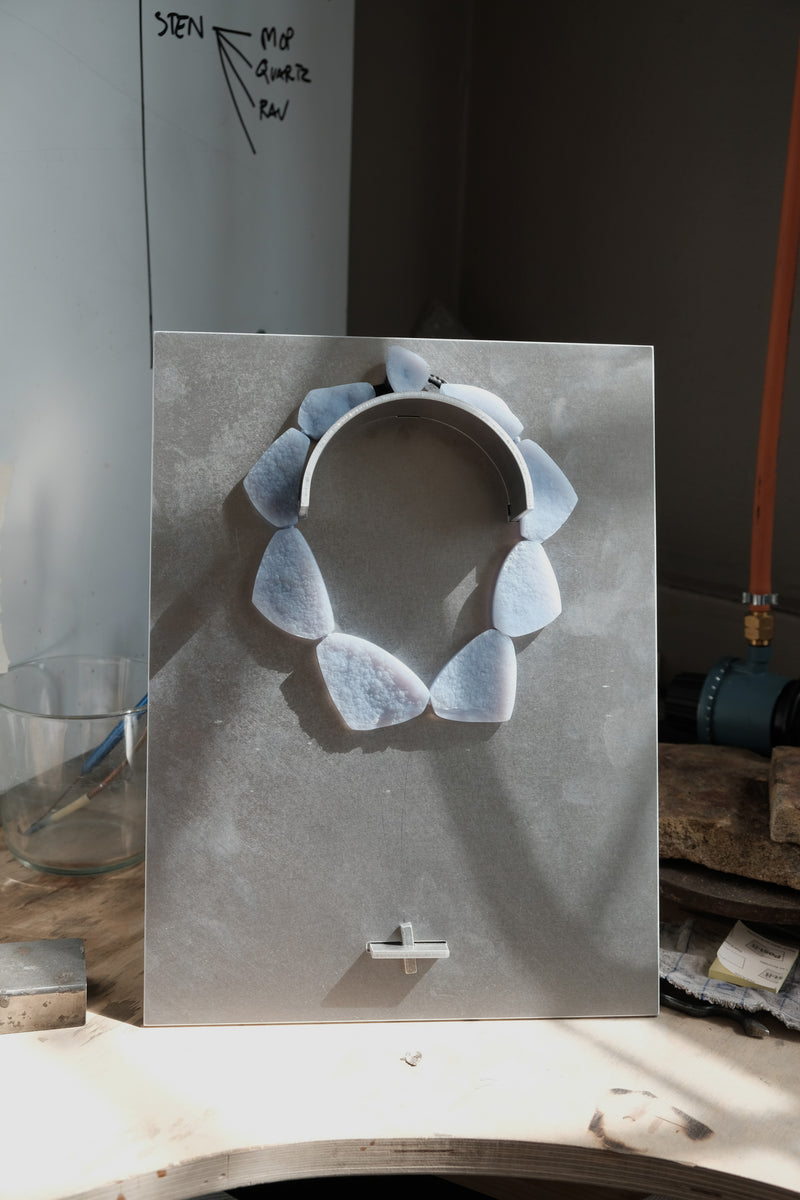 A handmade necklace made of irregular, light blue-gray stone pieces arranged in a circle on a white display card, set against a workshop background.