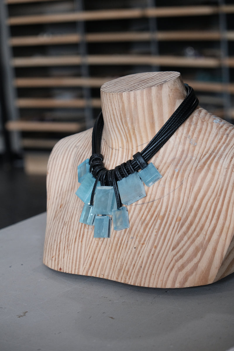 A statement necklace with light blue rectangular glass pendants and a black braided cord, displayed on a light wood mannequin bust.