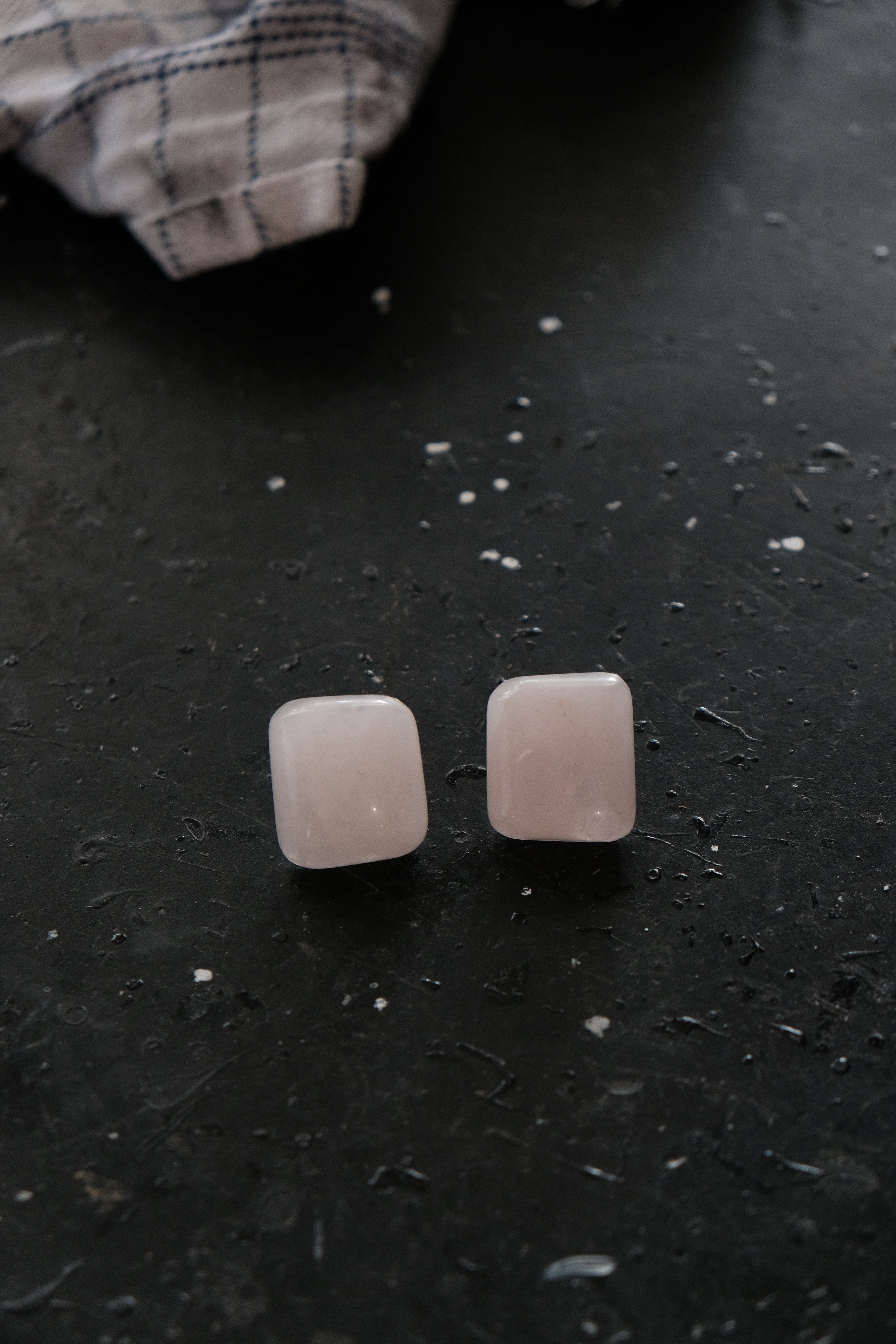A pair of square-shaped rose quartz stud earrings resting on a dark, textured surface with a blurred plaid cloth in the background.