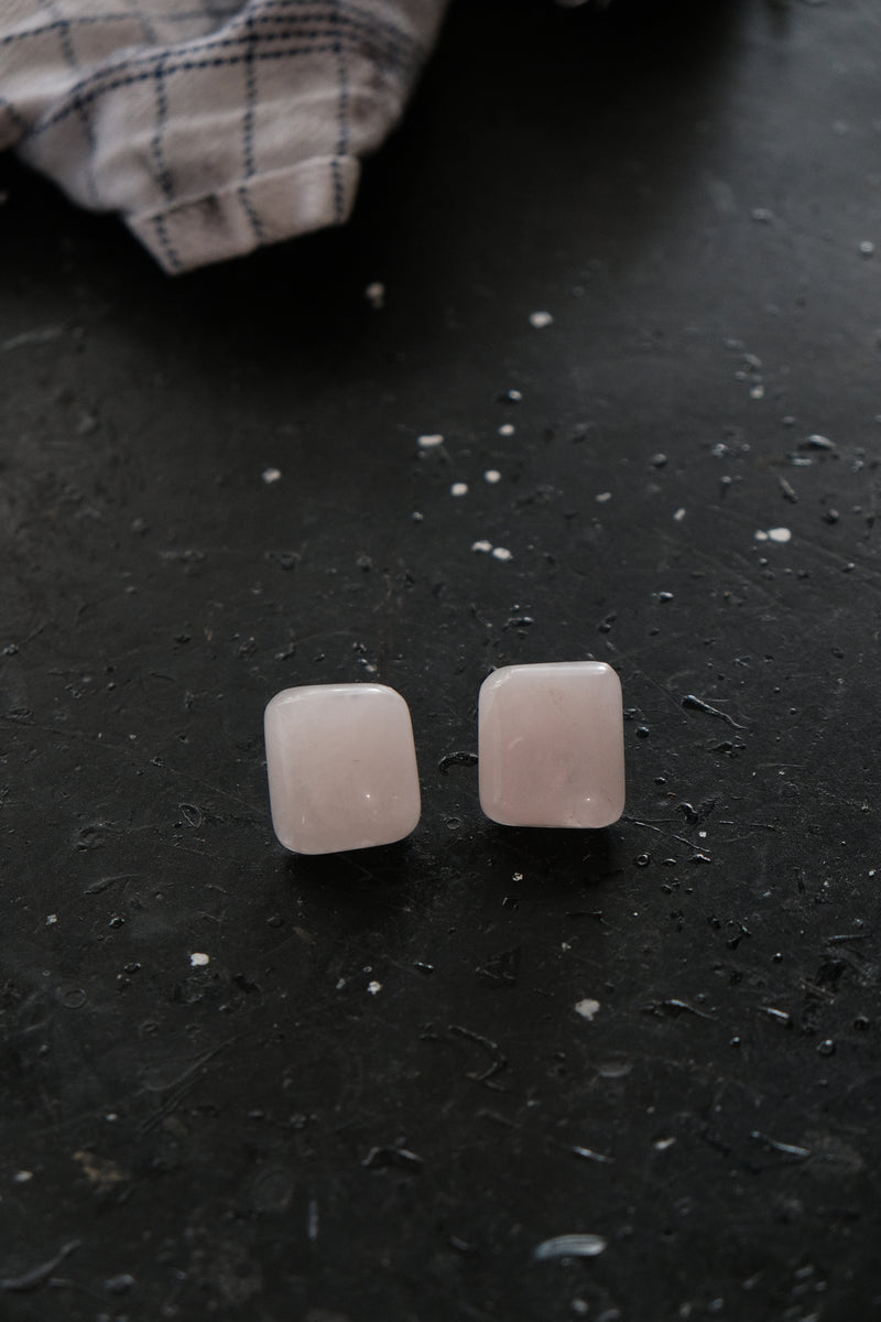 A pair of square-shaped rose quartz stud earrings resting on a dark, textured surface with a blurred plaid cloth in the background.