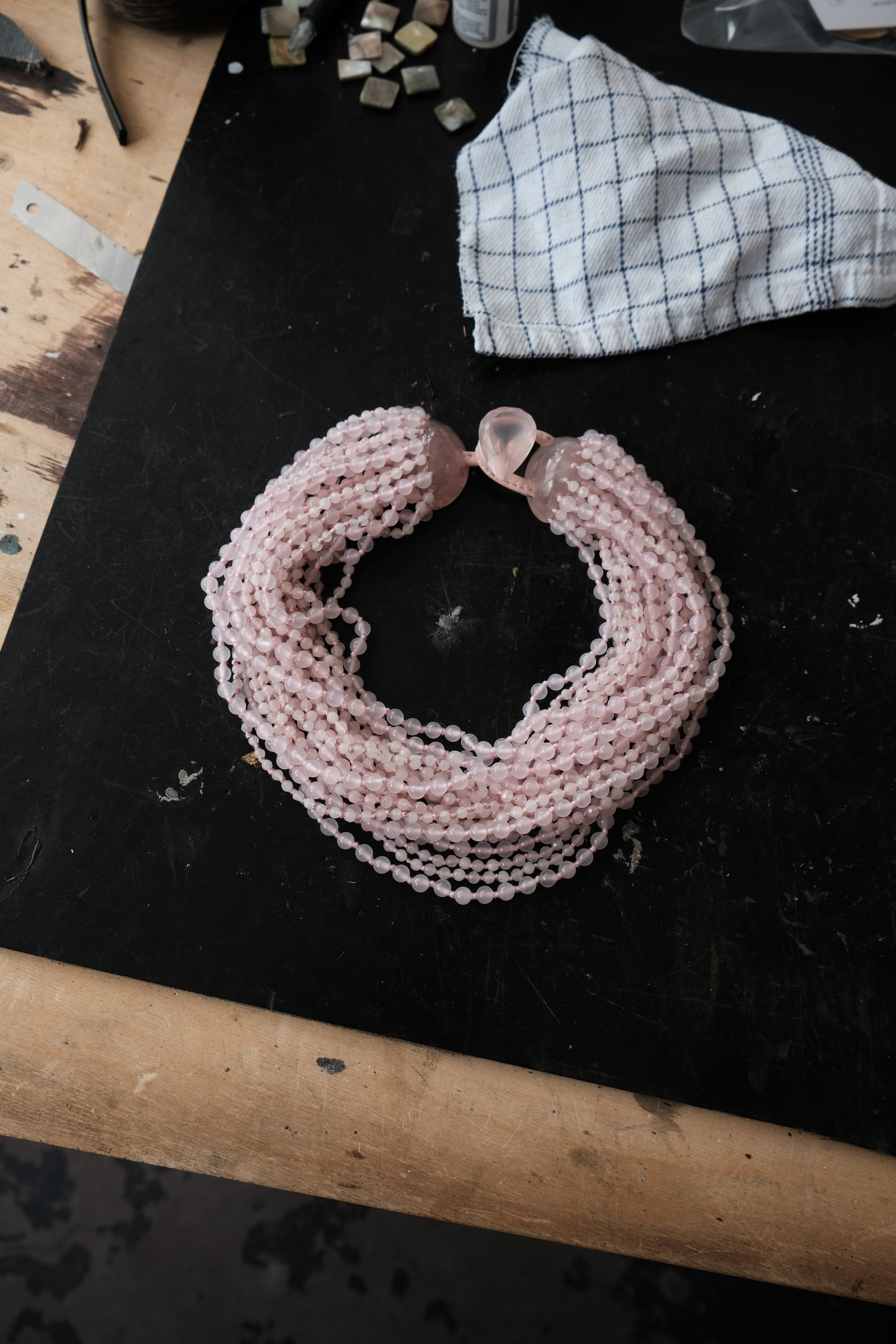 A multi-strand pink beaded necklace with a faceted pink stone clasp, displayed on a black workbench next to a checkered cloth.