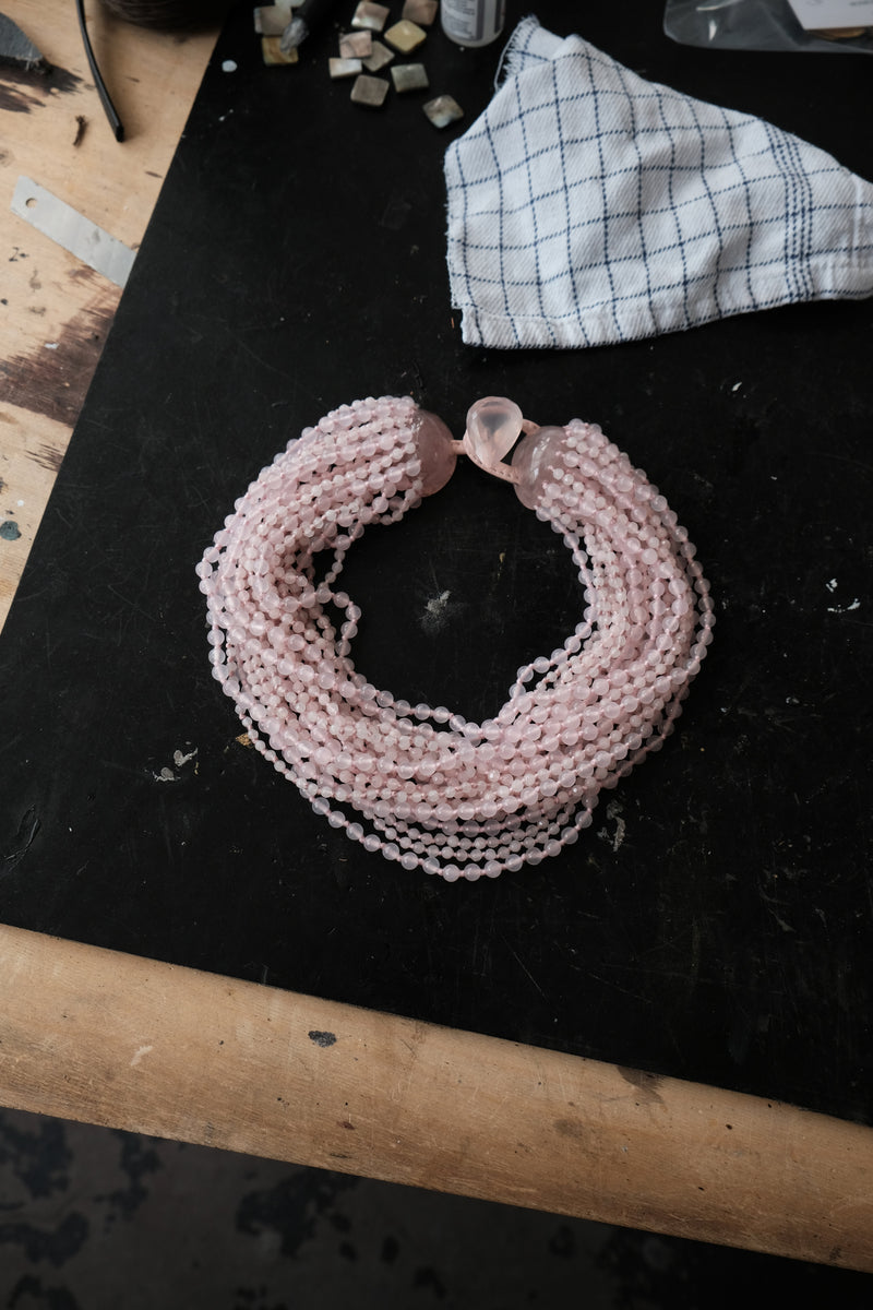 A multi-strand pink beaded necklace with a faceted pink stone clasp, displayed on a black workbench next to a checkered cloth.