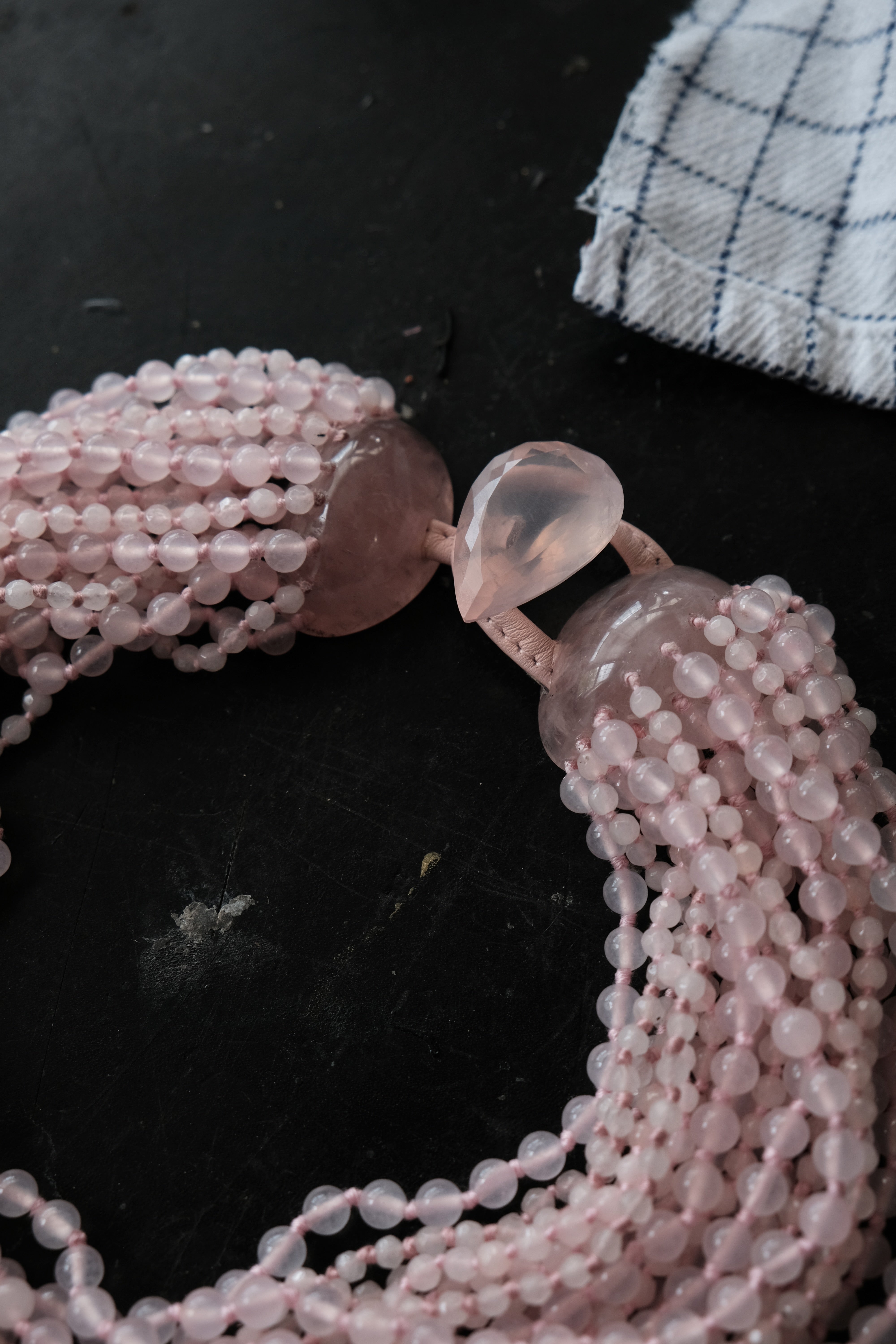 A close-up of a multi-strand rose quartz beaded necklace with polished teardrop-shaped beads, arranged on a dark surface with a small white and navy checkered cloth in the corner.