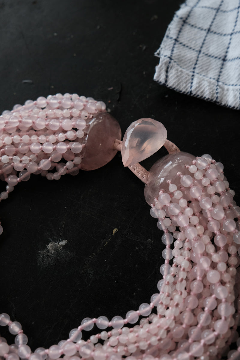 A close-up of a multi-strand rose quartz beaded necklace with polished teardrop-shaped beads, arranged on a dark surface with a small white and navy checkered cloth in the corner.