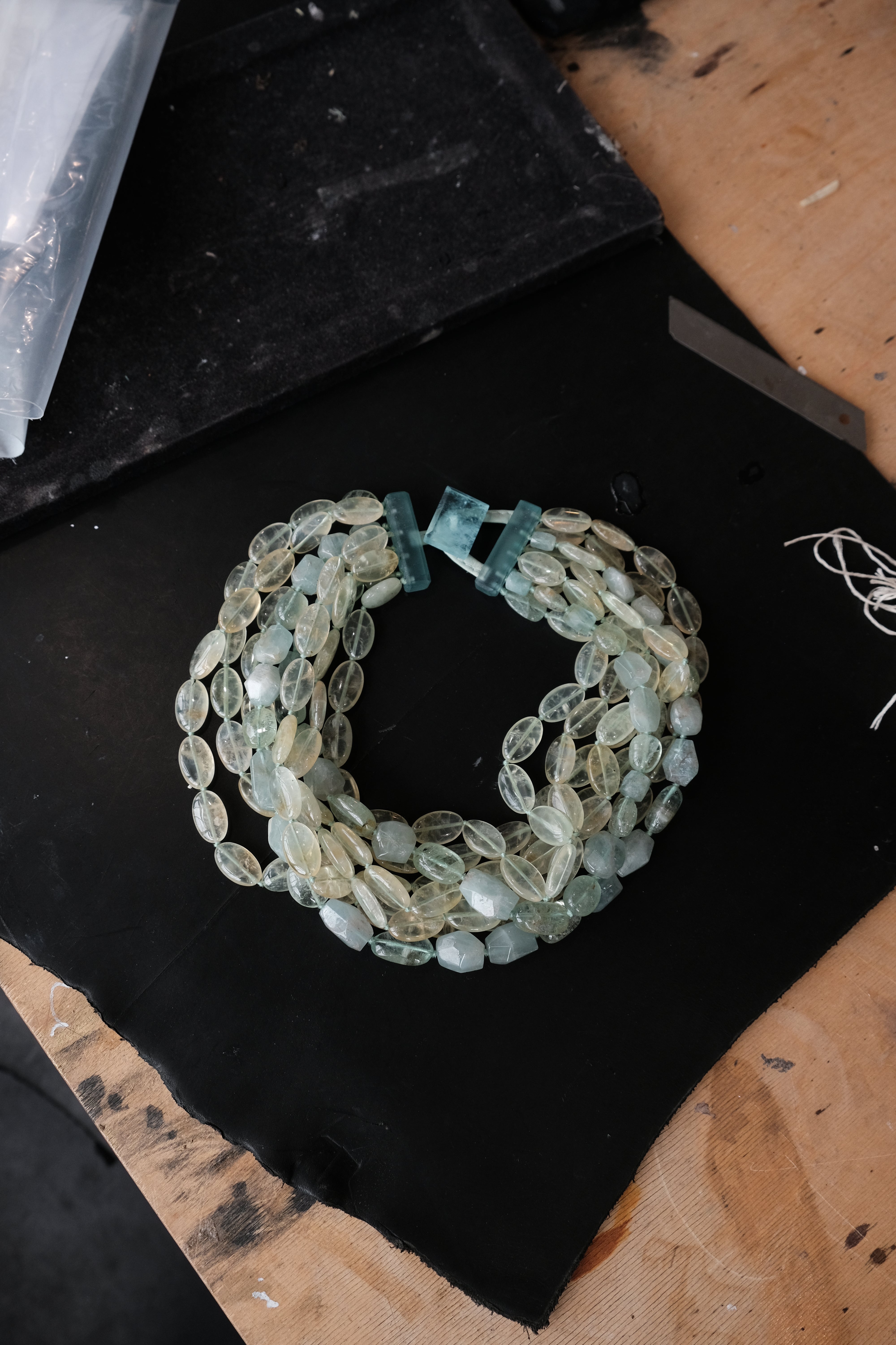 A handmade beaded necklace made of multiple strands of oval beads in shades of sage green, light blue, and cream, featuring a clasp of larger blue-green beads, displayed on a black surface.