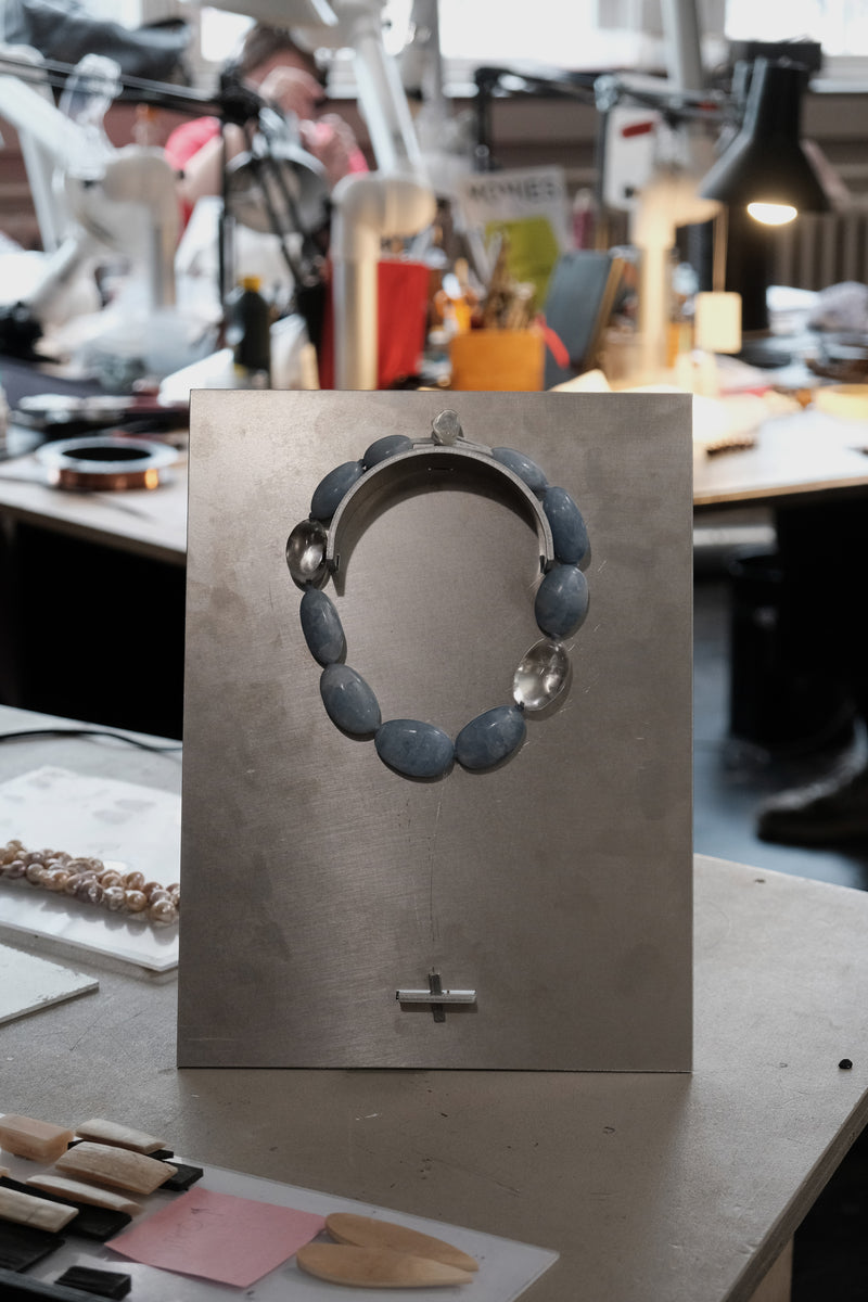 A handmade beaded necklace with slate blue oval beads and clear faceted accent beads, displayed on a metal stand in a workshop setting.