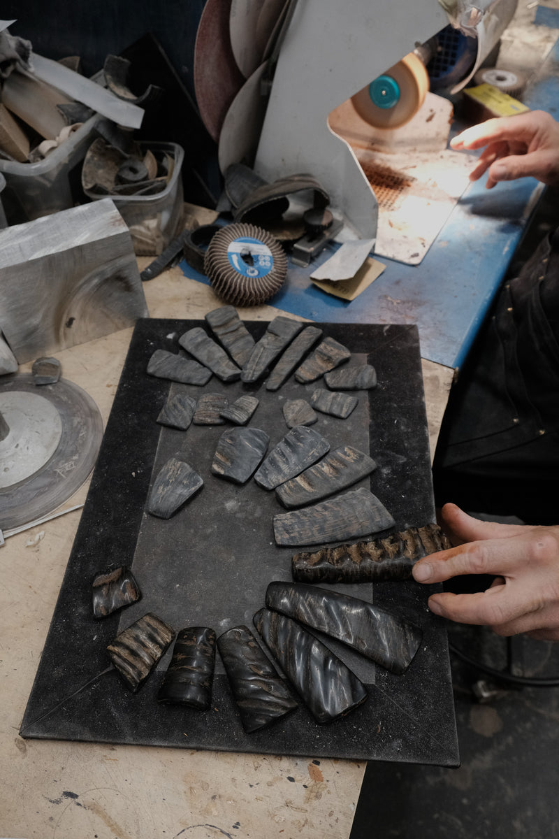 A workshop scene showing a black rectangular board with multiple dark metallic or stone-like rectangular pieces arranged around it, plus a person's hands holding one piece near a grinding machine.