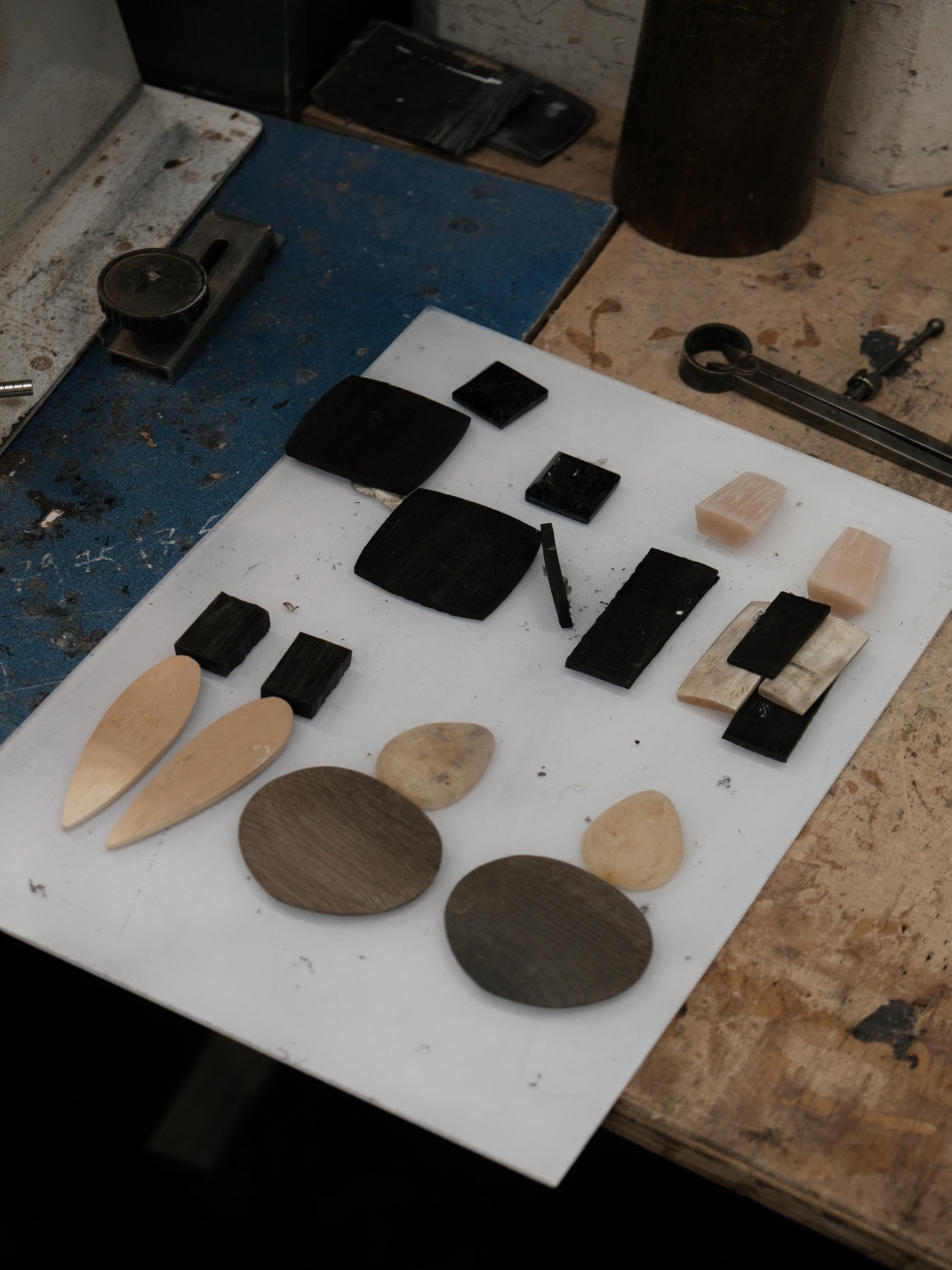 A collection of geometric horn and stone jewelry components, including oval, rectangular, and teardrop-shaped pieces in natural wood and dark stone, arranged on a white sheet on a workbench.