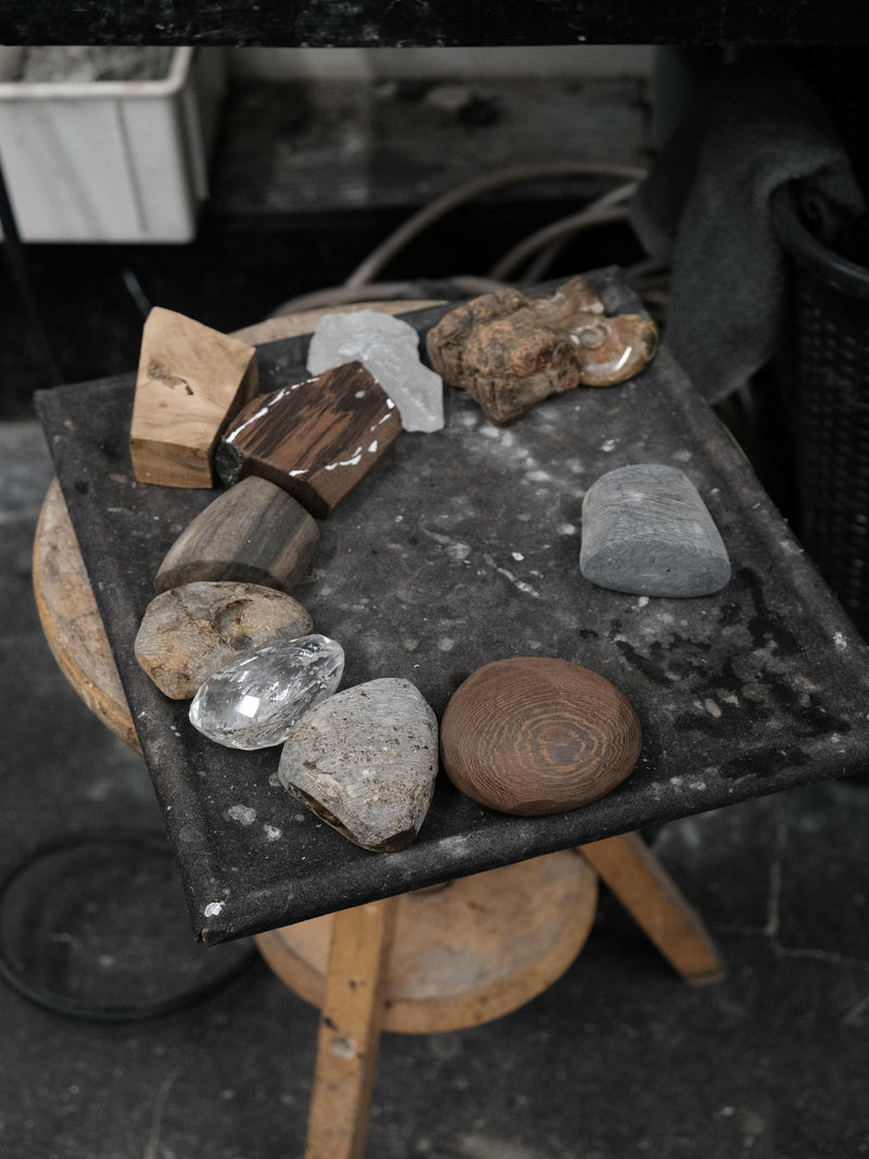 A collection of polished stones, crystals, and wood blocks arranged on a dark square tray resting on a wooden stool in a workshop setting.