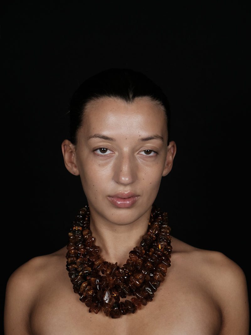 A close-up portrait of a model with dark hair pulled back, wearing a chunky multi-strand amber necklace against a solid black background.