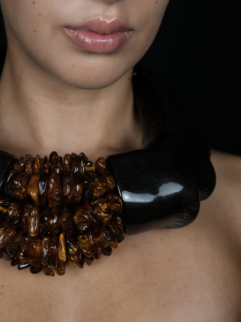 A close-up of a model wearing a chunky collar necklace made of polished amber-colored beads on one side and a glossy black wooden-like cylinder on the other, set against a dark background.