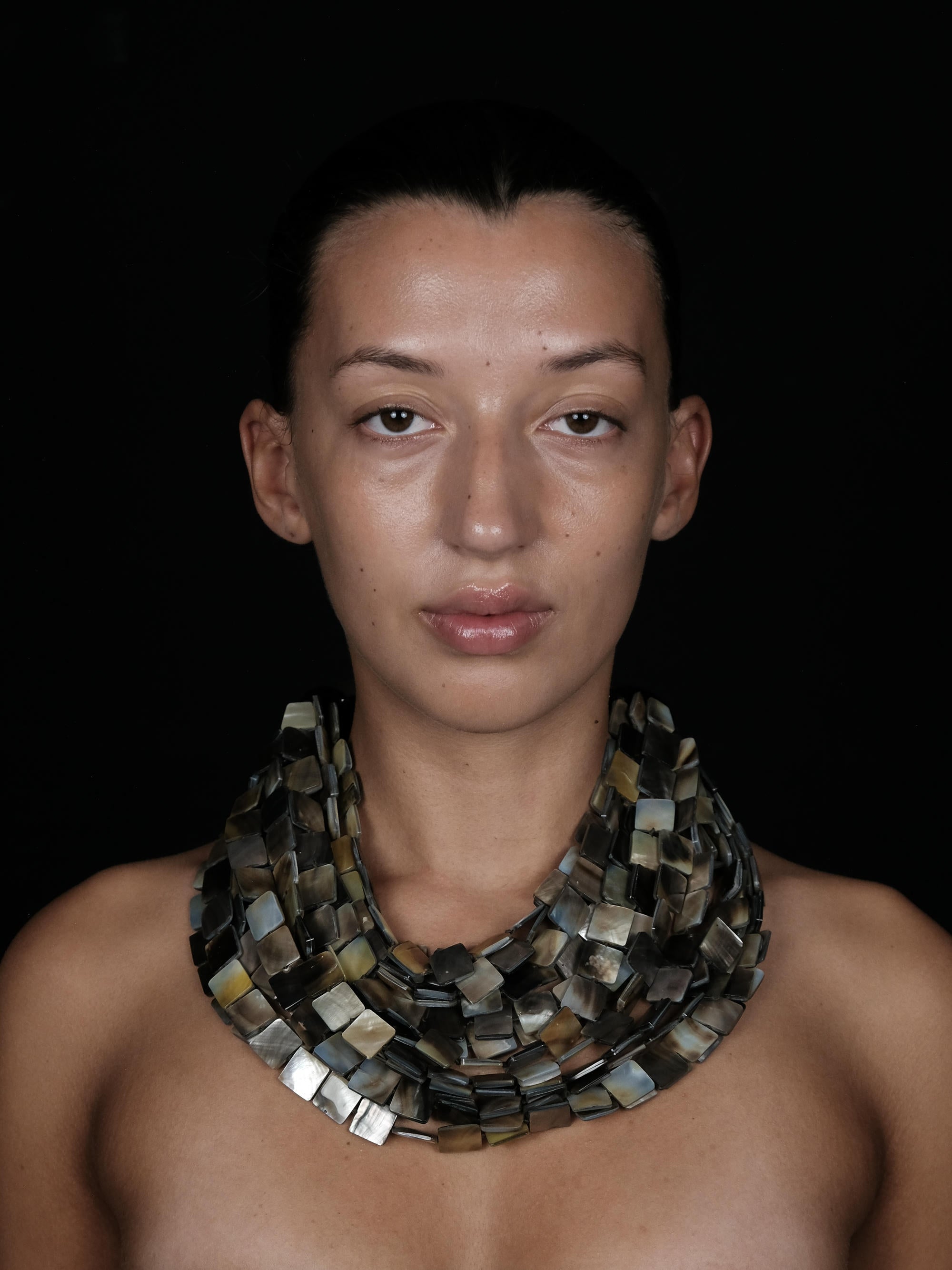 A close-up portrait of a model wearing a chunky, multi-strand collar necklace made of rectangular mother-of-pearl tiles in earthy tones of gray, beige, and brown, set against a solid black background.