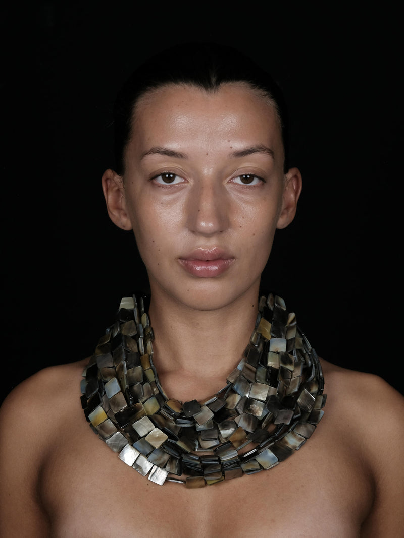 A close-up portrait of a model wearing a chunky, multi-strand collar necklace made of rectangular mother-of-pearl tiles in earthy tones of gray, beige, and brown, set against a solid black background.