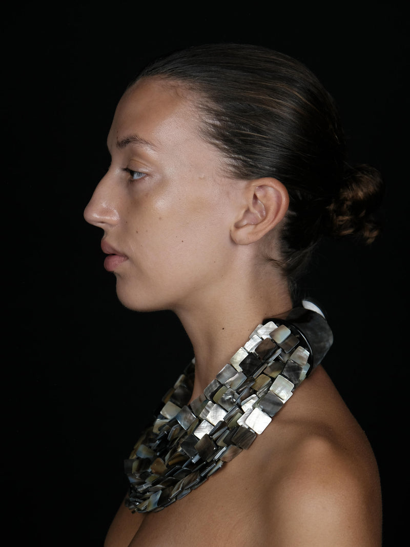 A side-profile portrait of a model wearing a chunky, geometric silver necklace made of overlapping square tiles, set against a solid black background.