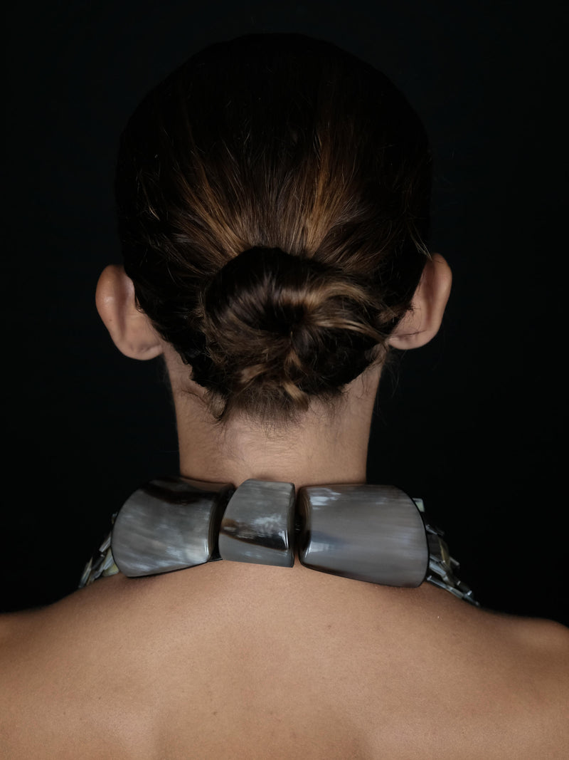 Rear view of a person with dark hair in a low bun wearing a chunky collar made of large polished gray stone-like segments, against a black background.