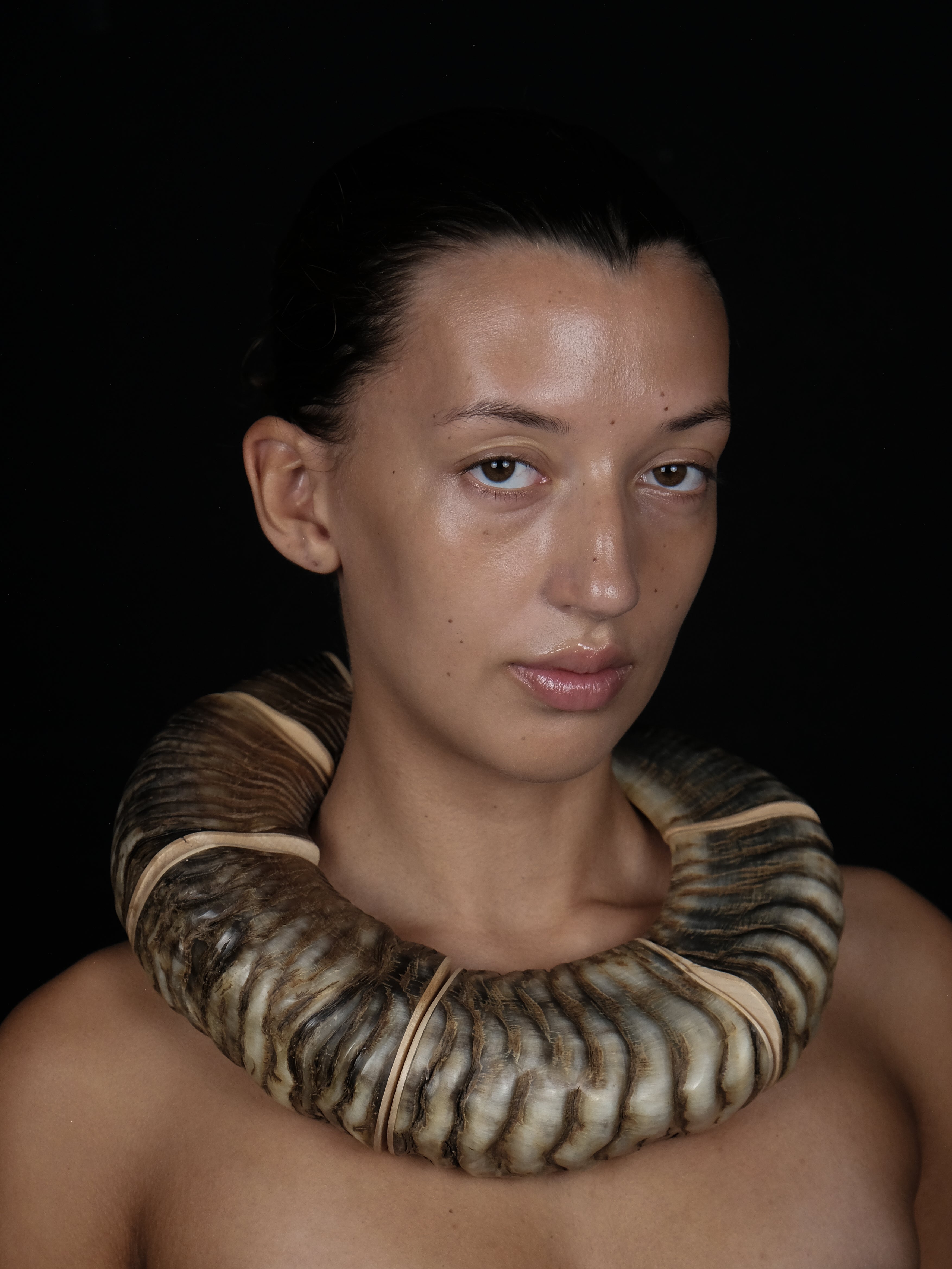 A close-up portrait of a person wearing a large, thick, ribbed horn necklace against a solid black background.