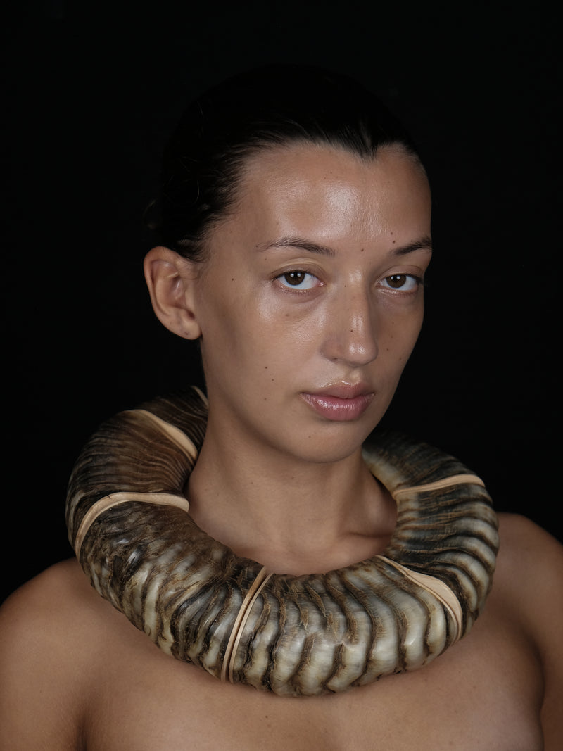 A close-up portrait of a person wearing a large, thick, ribbed horn necklace against a solid black background.