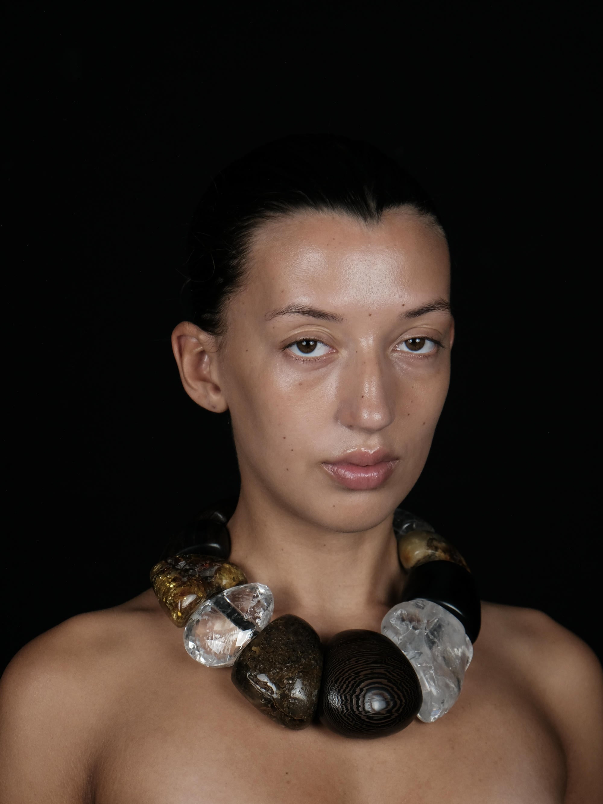 A close-up portrait of a model wearing a chunky statement necklace made of large polished stones and clear crystal-like beads, set against a solid black background.