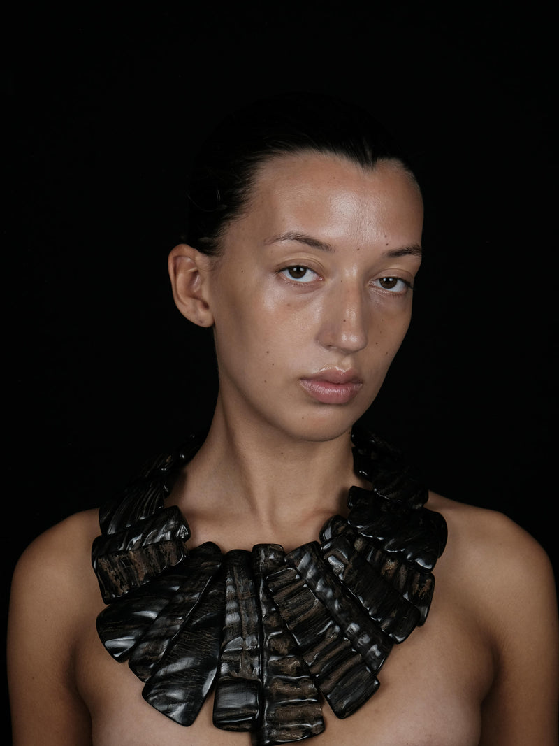 A model with dark hair pulled back wears a large, sculptural black collar necklace made of layered, textured leaf-like pieces against a solid black background.