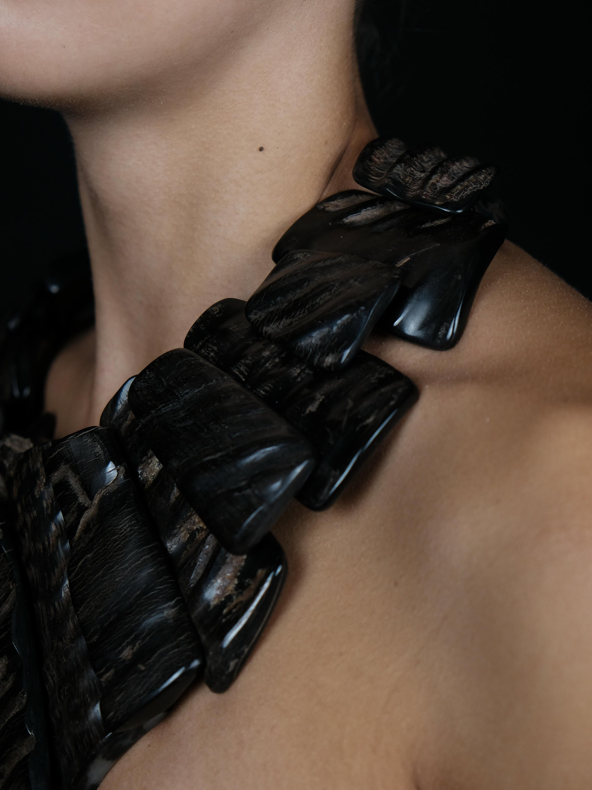 A close-up of a model's neck and shoulder wearing a chunky, sculptural black collar made of layered, textured petal-like or shell-like pieces.
