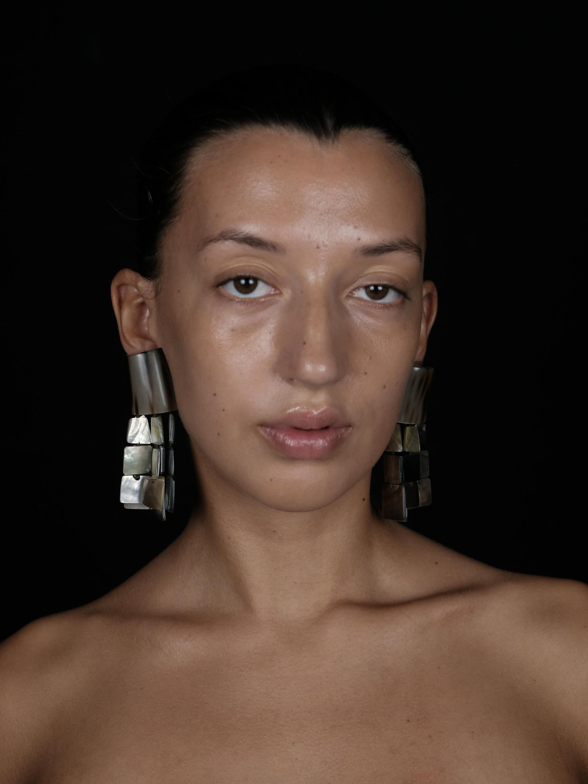 A close-up portrait of a model wearing large, chunky geometric silver earrings against a solid black background.