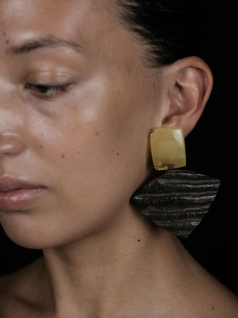 A close-up of a model wearing a large, dual-texture statement earring. The earring features a translucent, warm yellow top section and a dark, textured, charcoal-gray bottom section with horizontal striations, set against a solid black background.