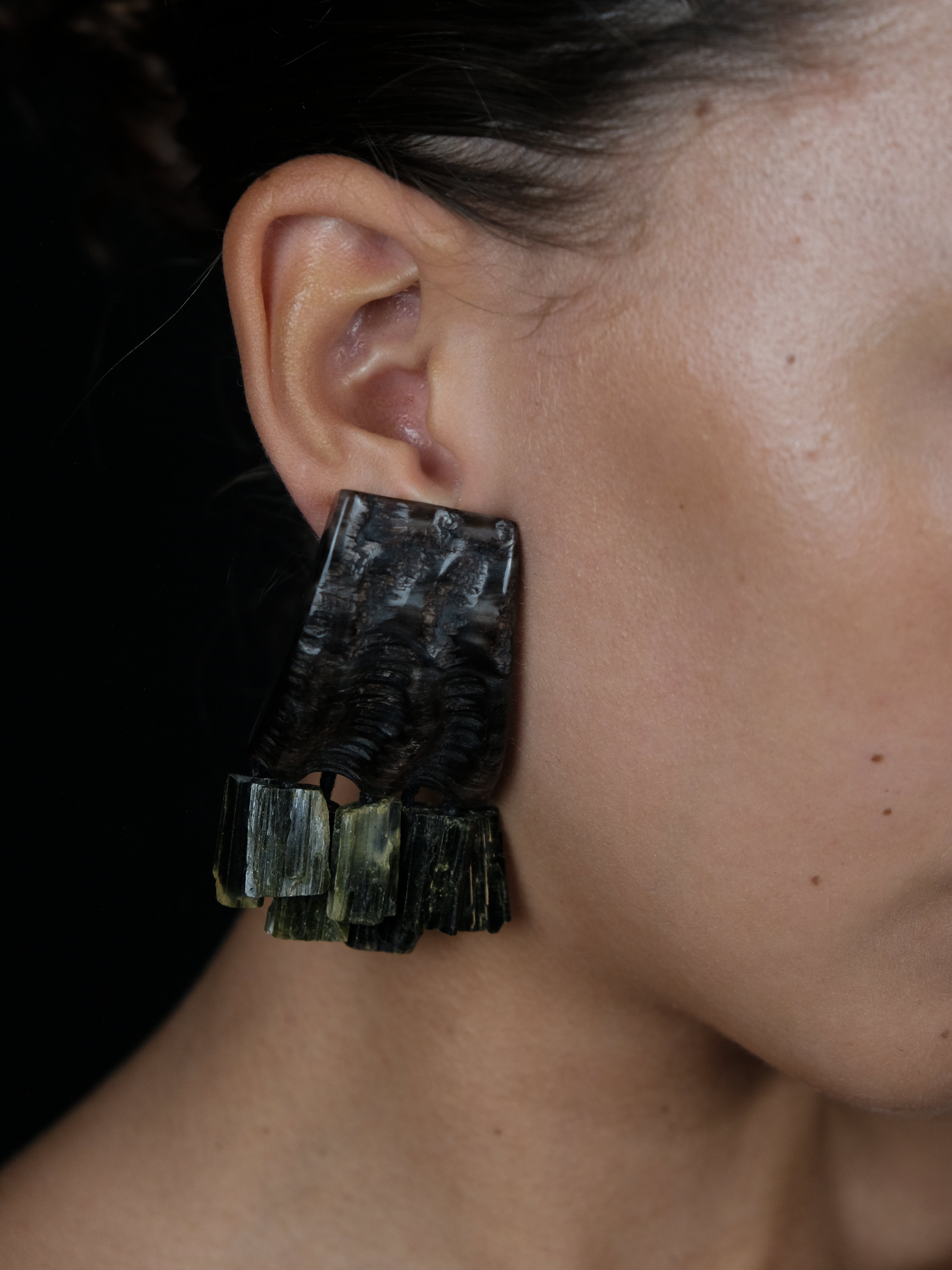 A close-up of a person's ear wearing a large, textured black earring with layered rectangular stone-like drops in dark green and black.