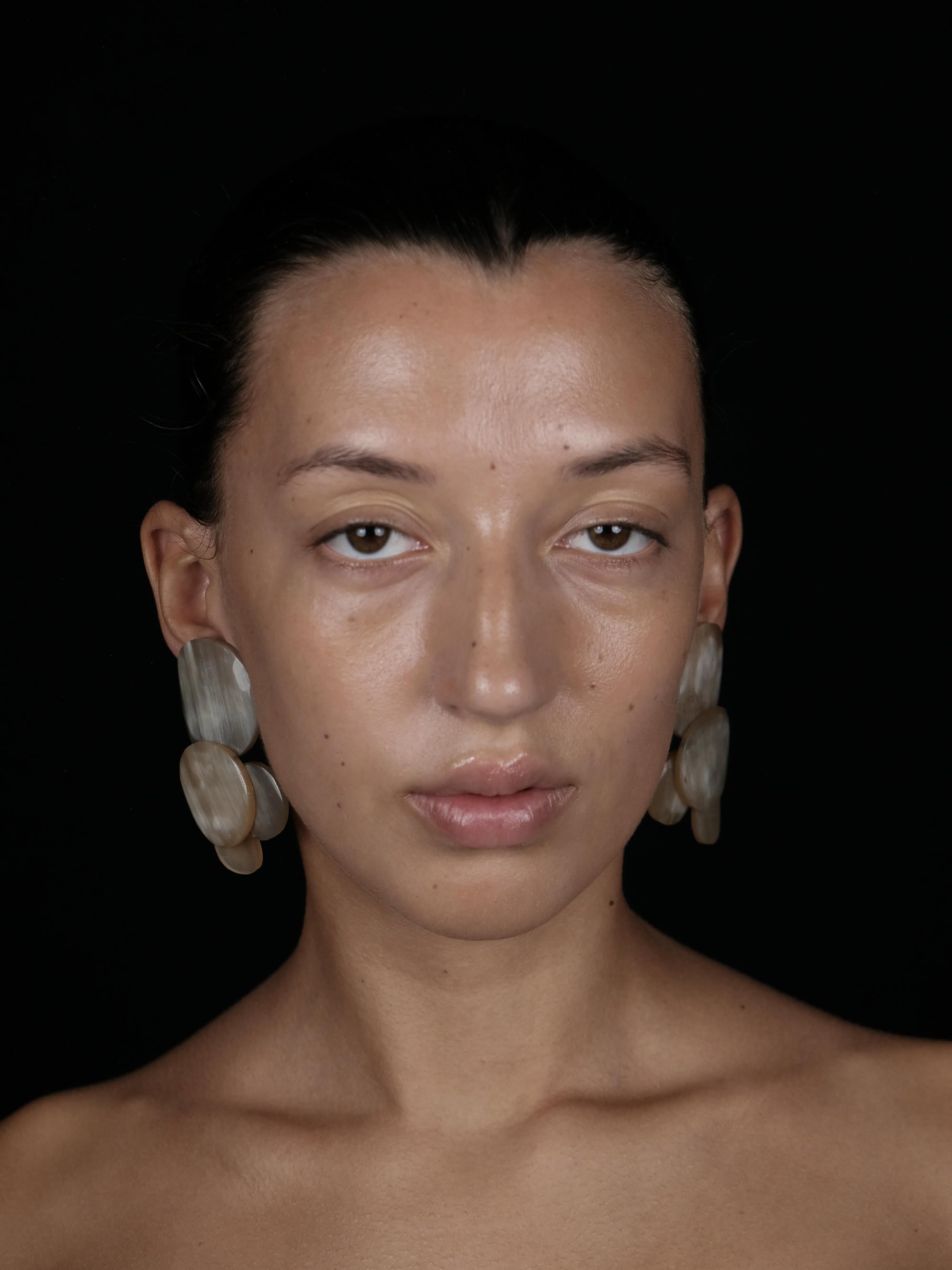 A close-up portrait of a model with dark hair pulled back, wearing large, stacked, disc-like shell earrings against a solid black background.