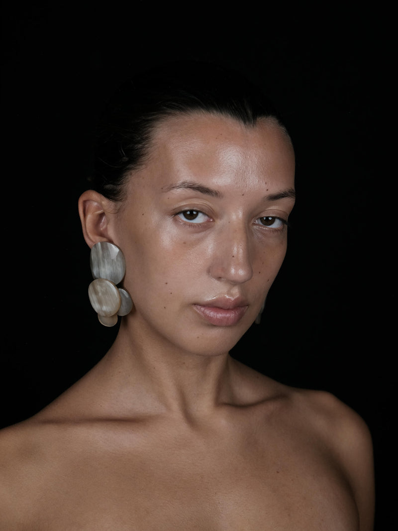 A close-up portrait of a woman with dark hair pulled back, wearing large layered shell earrings against a solid black background.