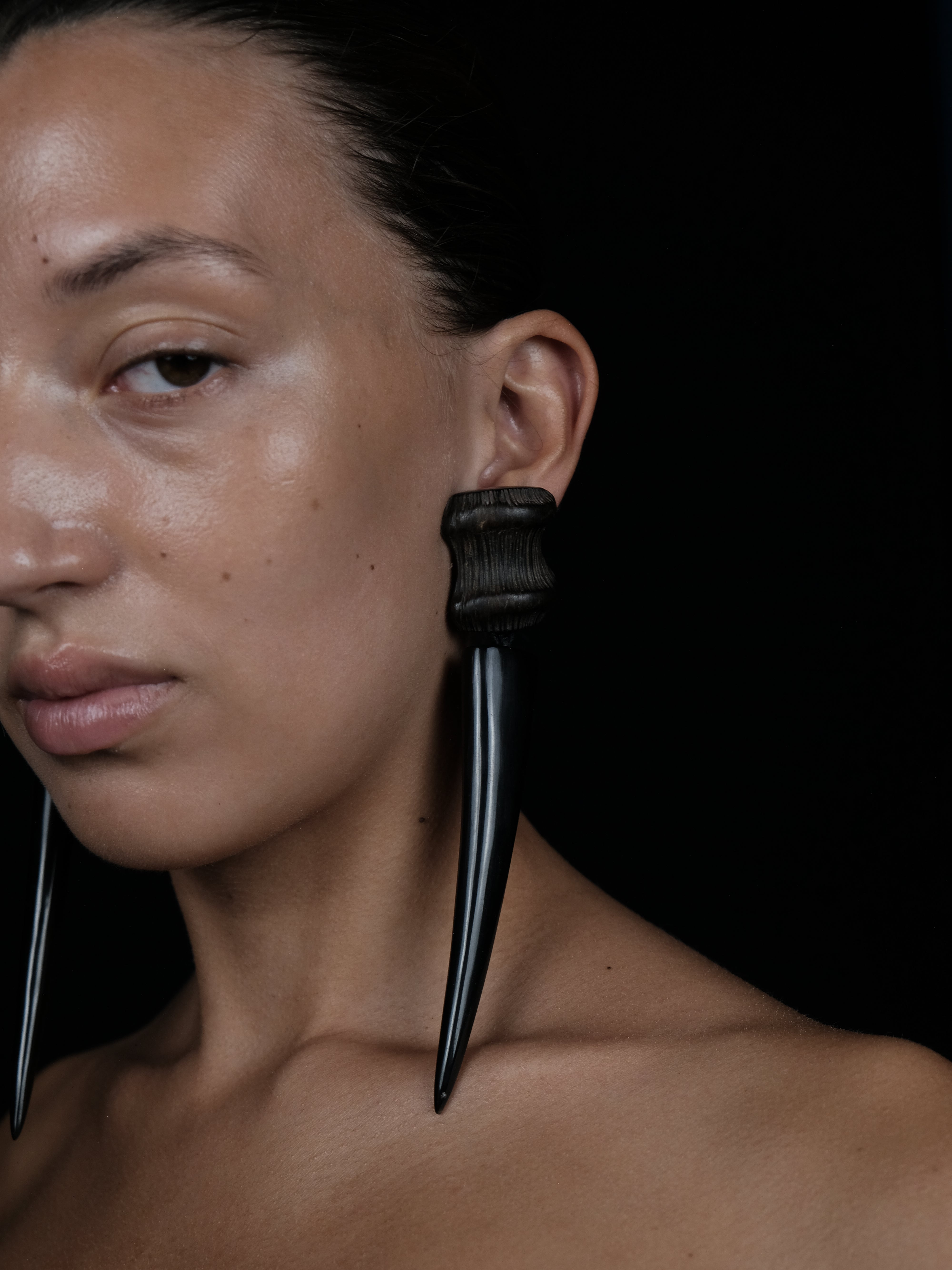 A close-up portrait of a model wearing a large black ear cuff with two long, curved, spike-like dangling elements, set against a solid black background.