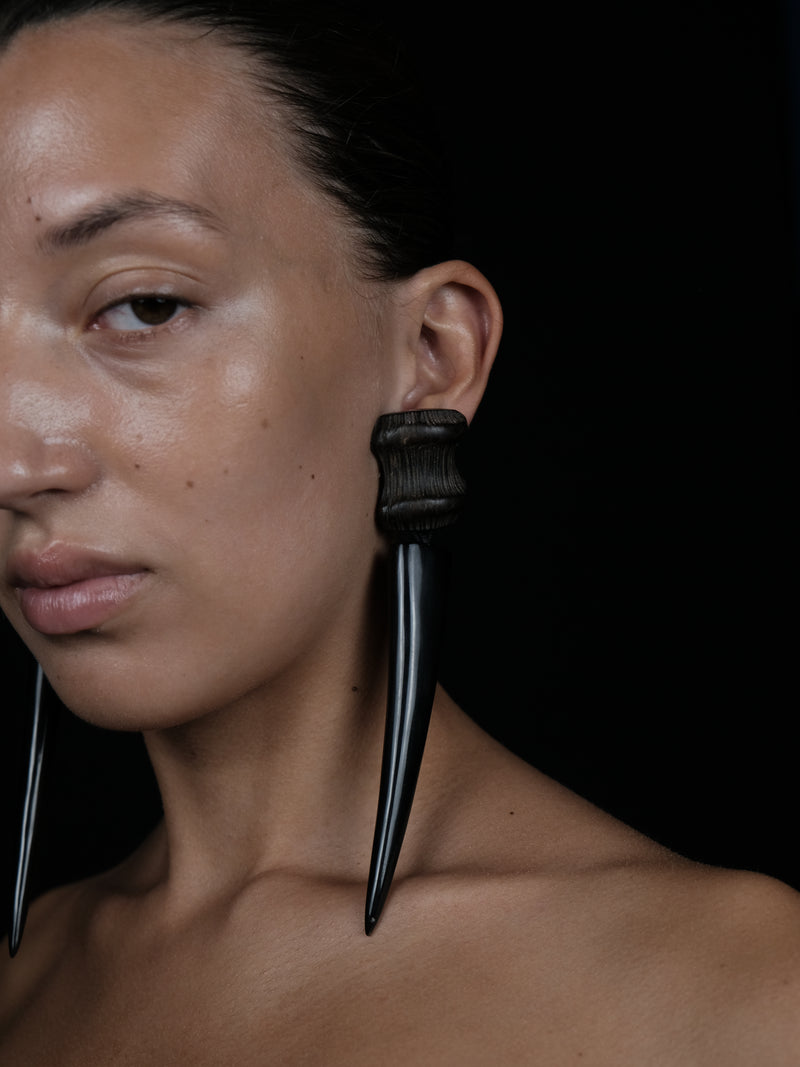 A close-up portrait of a model wearing a large black ear cuff with two long, curved, spike-like dangling elements, set against a solid black background.