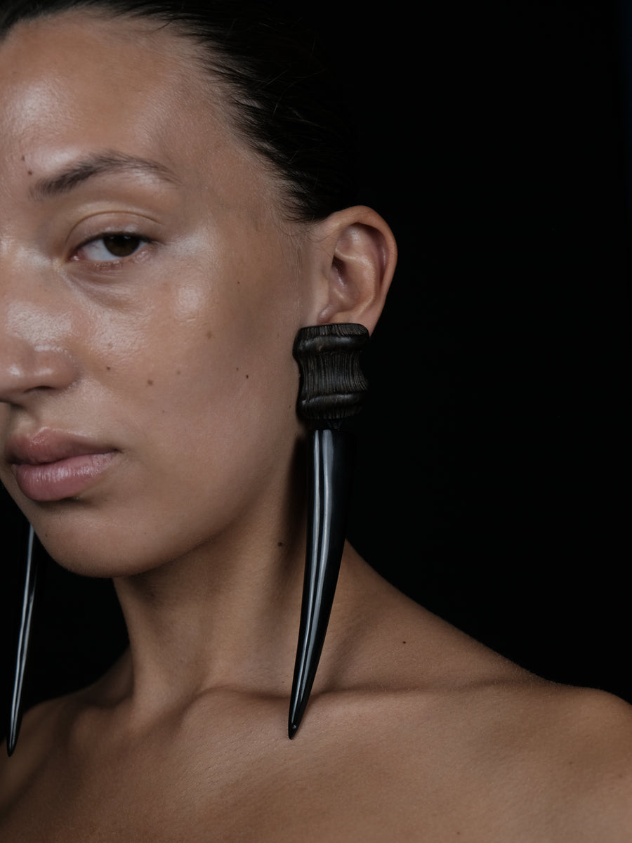A close-up portrait of a model wearing a large black ear cuff with two long, curved, spike-like dangling elements, set against a solid black background.