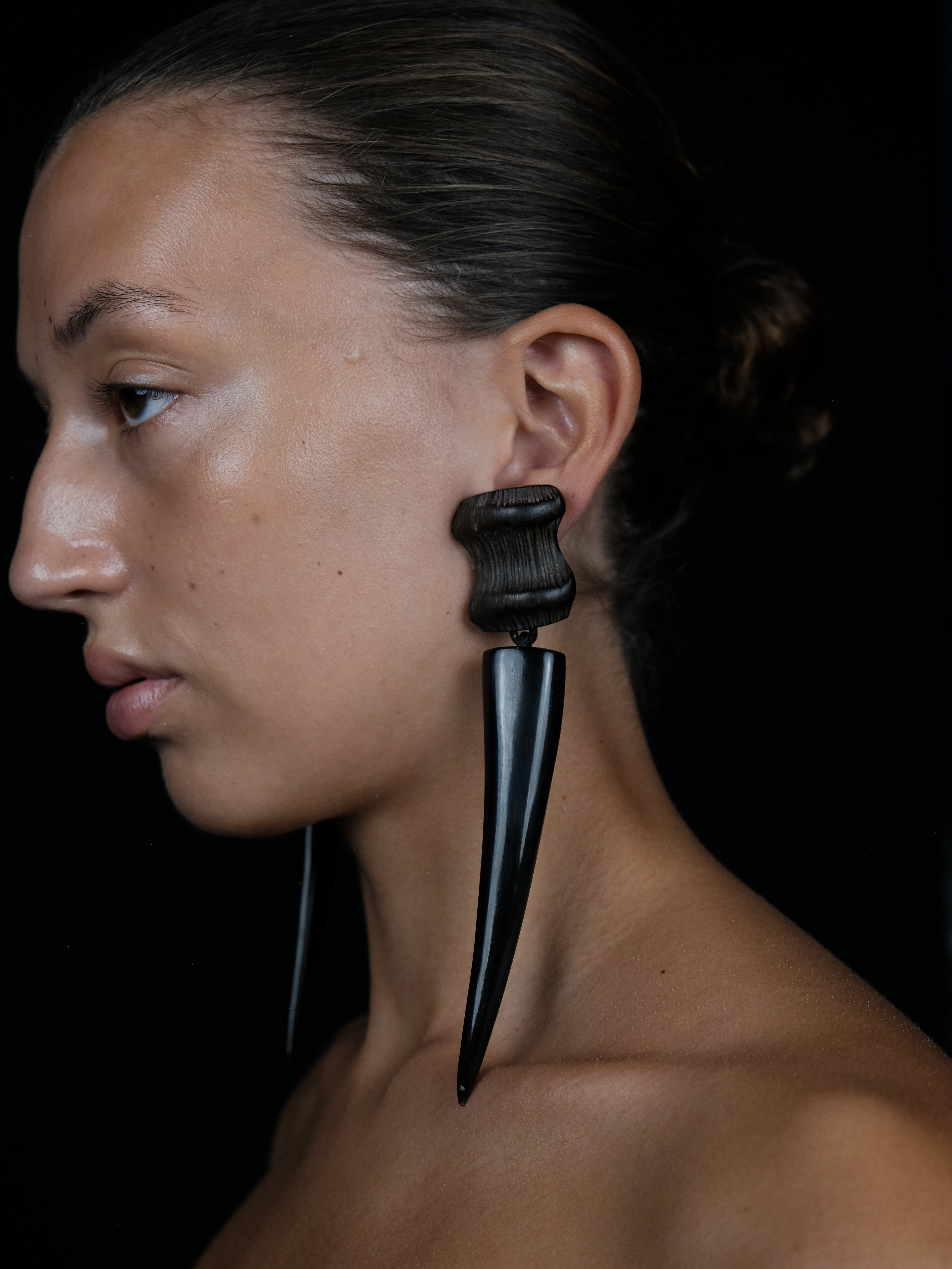 A close-up side profile of a model wearing a dramatic, large black dangle earring with a textured top and a tapered spike, set against a solid black background.