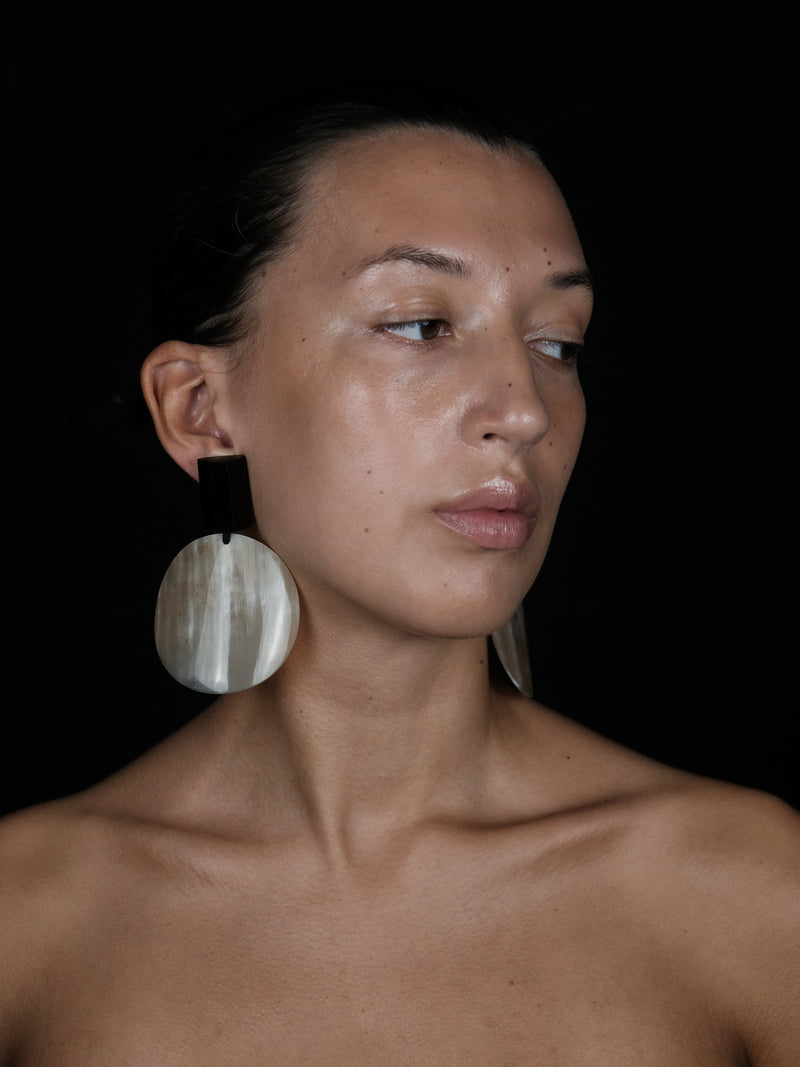 A close-up portrait of a woman wearing a large, circular, horn style earring with a black top tab, set against a solid black background.