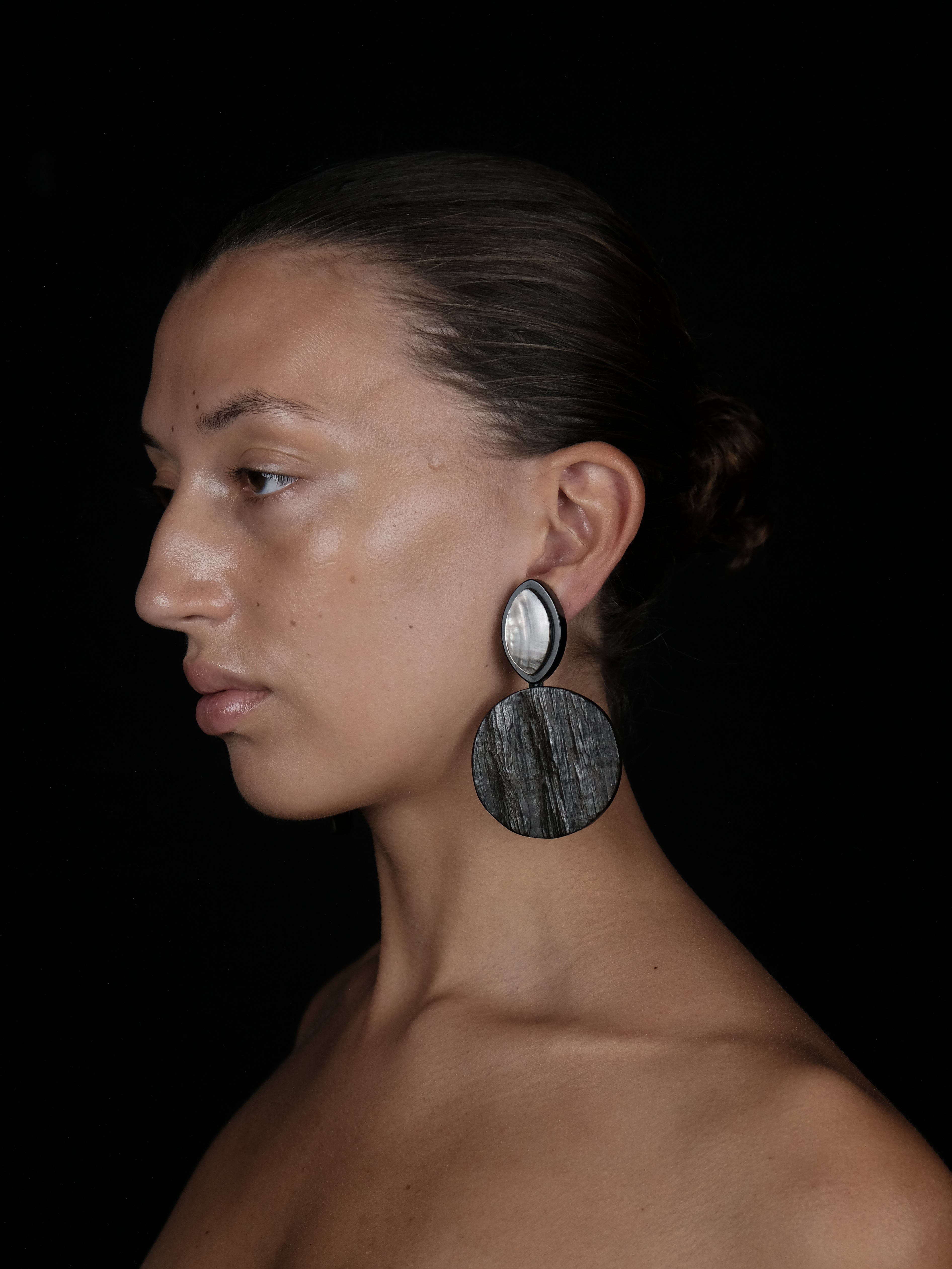 A close-up side profile of a woman wearing a large, circular drop earring with a silver-toned top disc and a dark, wood-grain textured bottom disc, set against a solid black background.