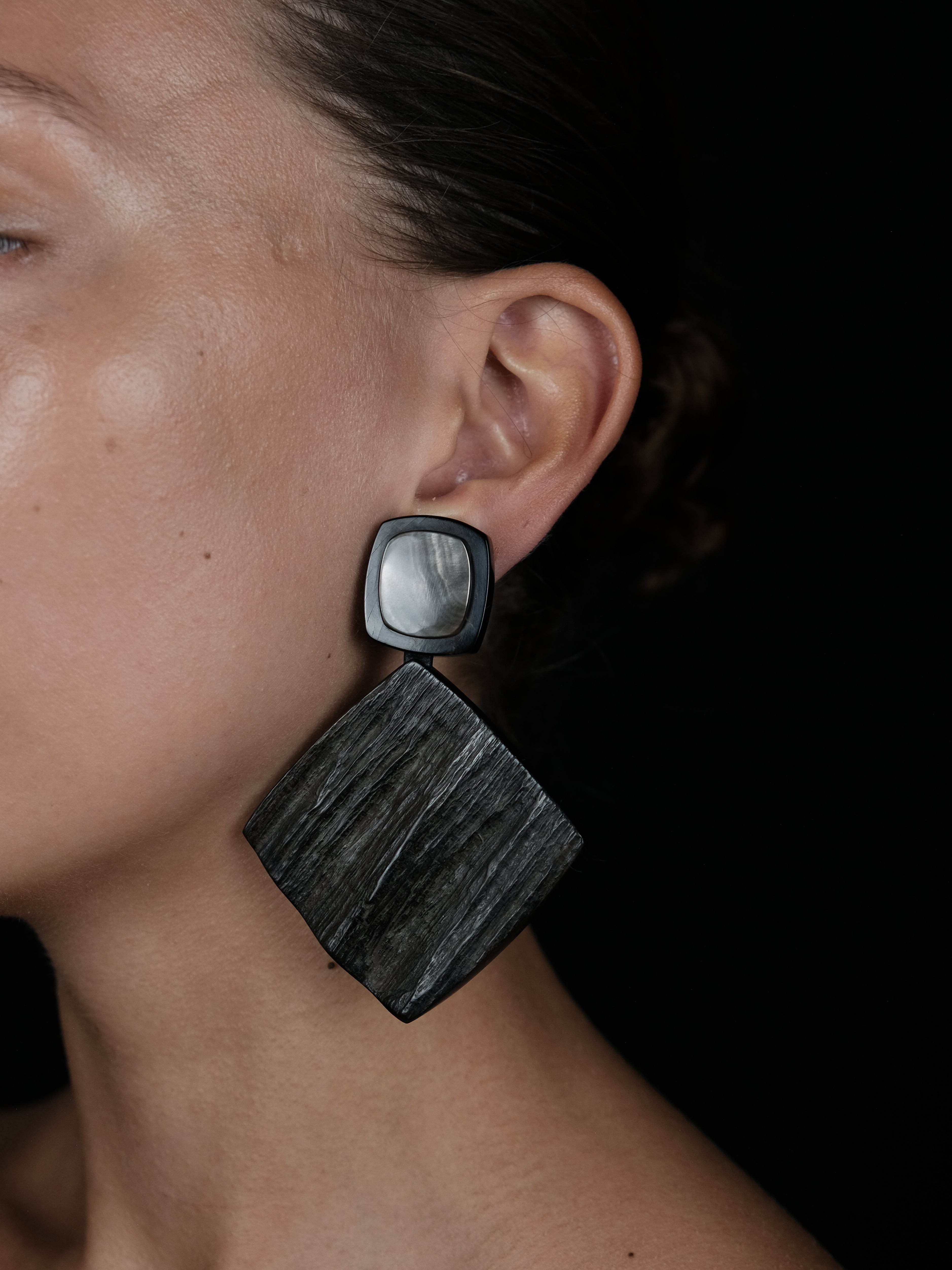 A close-up of a person's ear wearing a large geometric dangle earring. The earring features a square black top with a smooth, iridescent silver center, suspended from a larger rectangular black drop with a visible wood grain texture, set against a solid black background.