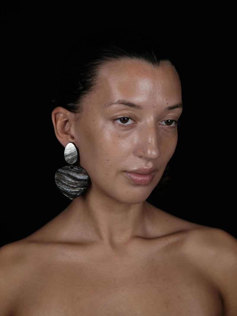 A close-up portrait of a woman with dark hair pulled back, wearing a large textured silver earring, set against a solid black background.