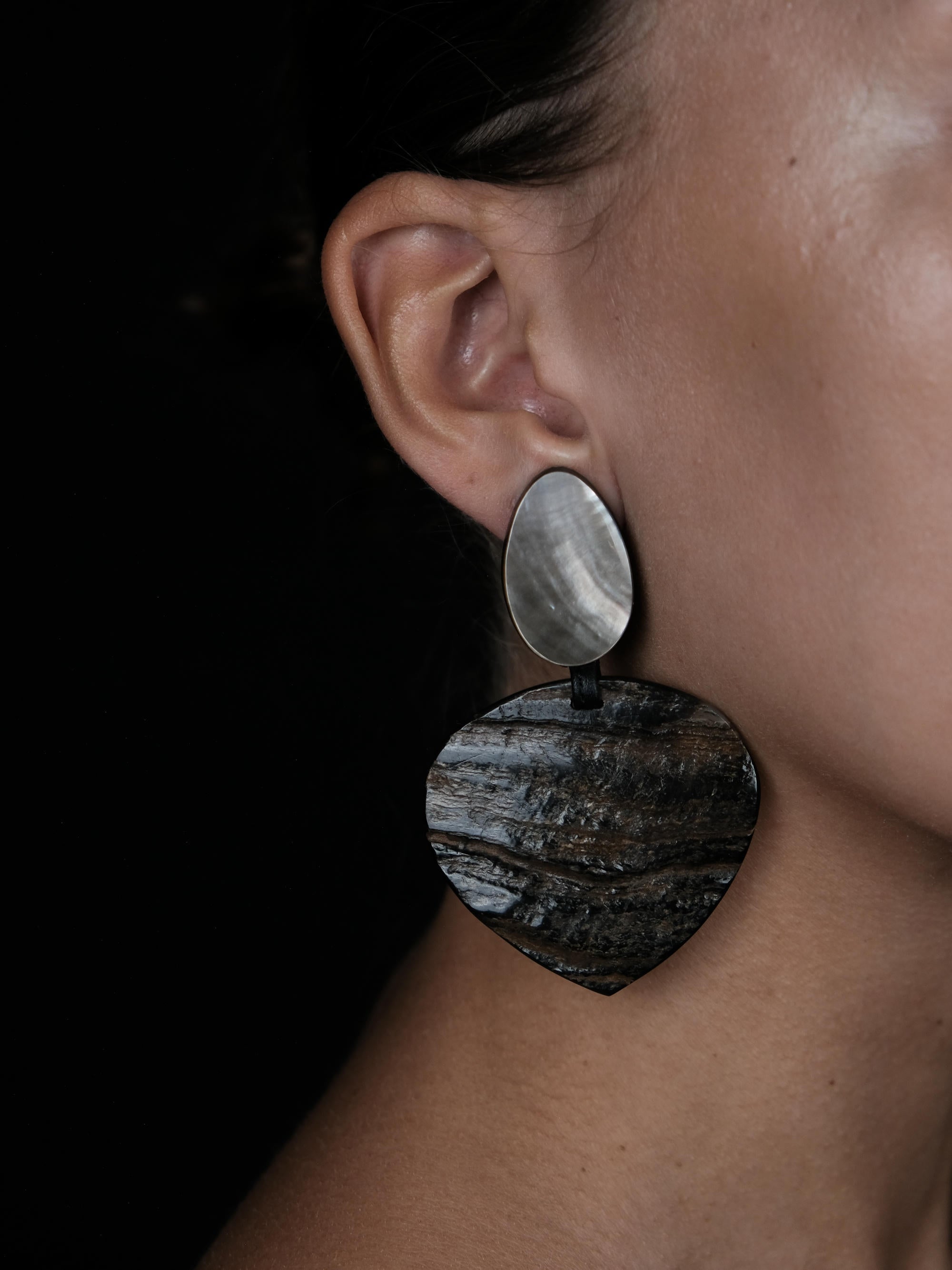 A close-up of a person's ear wearing a large statement earring. The earring has a smaller oval silver-toned top section and a larger irregular dark wood-textured drop section below, set against a dark background.