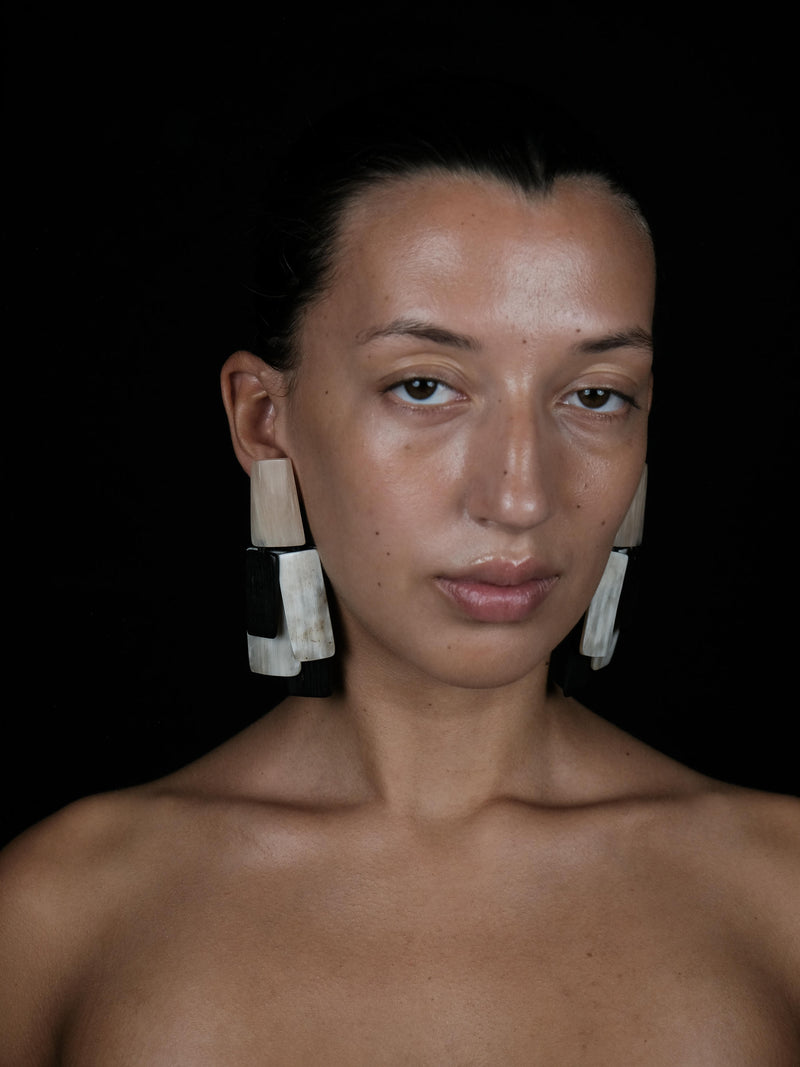 A close-up portrait of a woman with dark hair pulled back, wearing large geometric drop earrings made of stacked rectangular pieces in off-white, black, and beige tones, set against a solid black background.