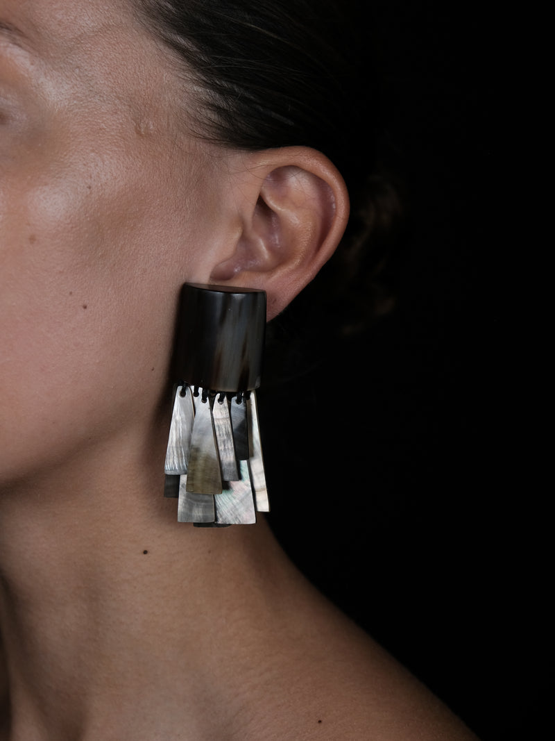 A close-up side view of a person's ear wearing a dramatic statement earring. The earring features a matte black top section and multiple layered, metallic silver and silver-gray fringe elements below, set against a dark background.