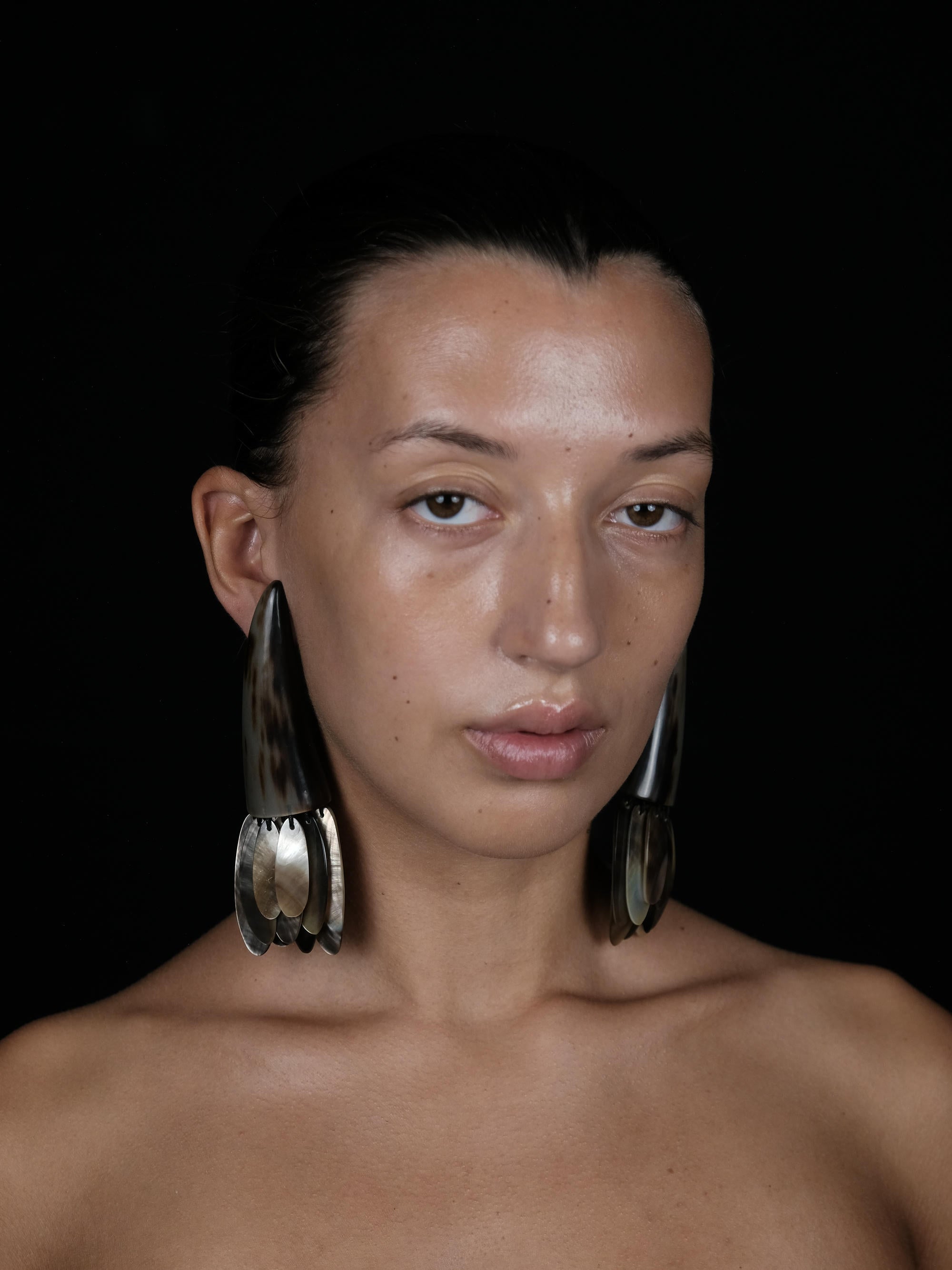 A close-up portrait of a model wearing large, sculptural silver-toned earrings against a solid black background.