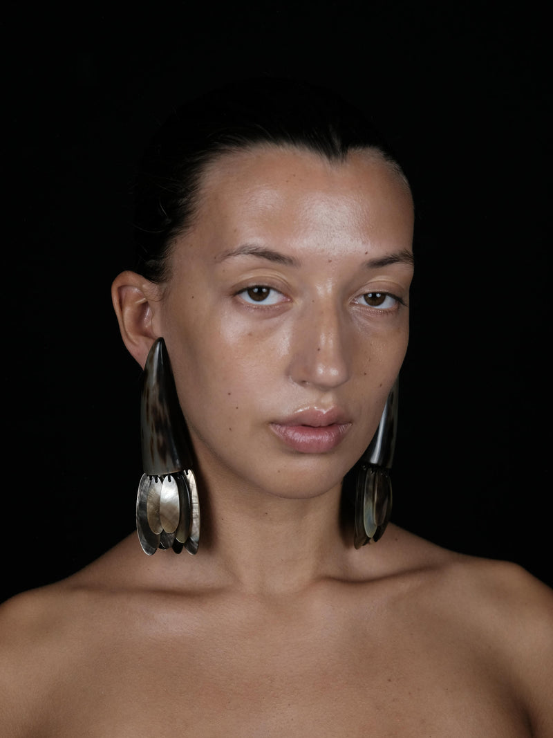A close-up portrait of a model wearing large, sculptural silver-toned earrings against a solid black background.